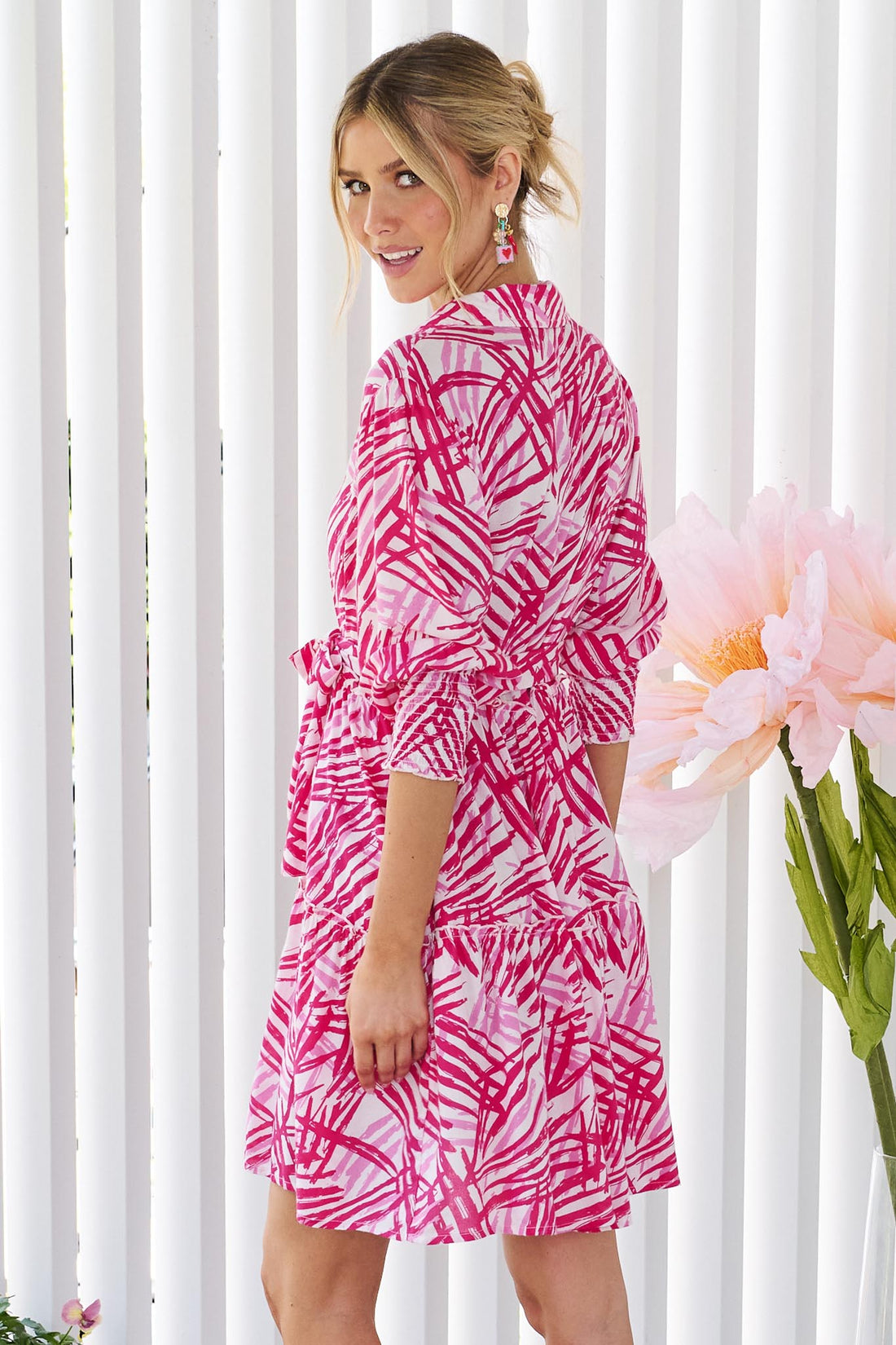 A woman wearing the minnie + MAXXIE Gemma pink and white abstract patterned mini dress with three-quarter sleeves and a tie waist, standing against a white panelled wall.