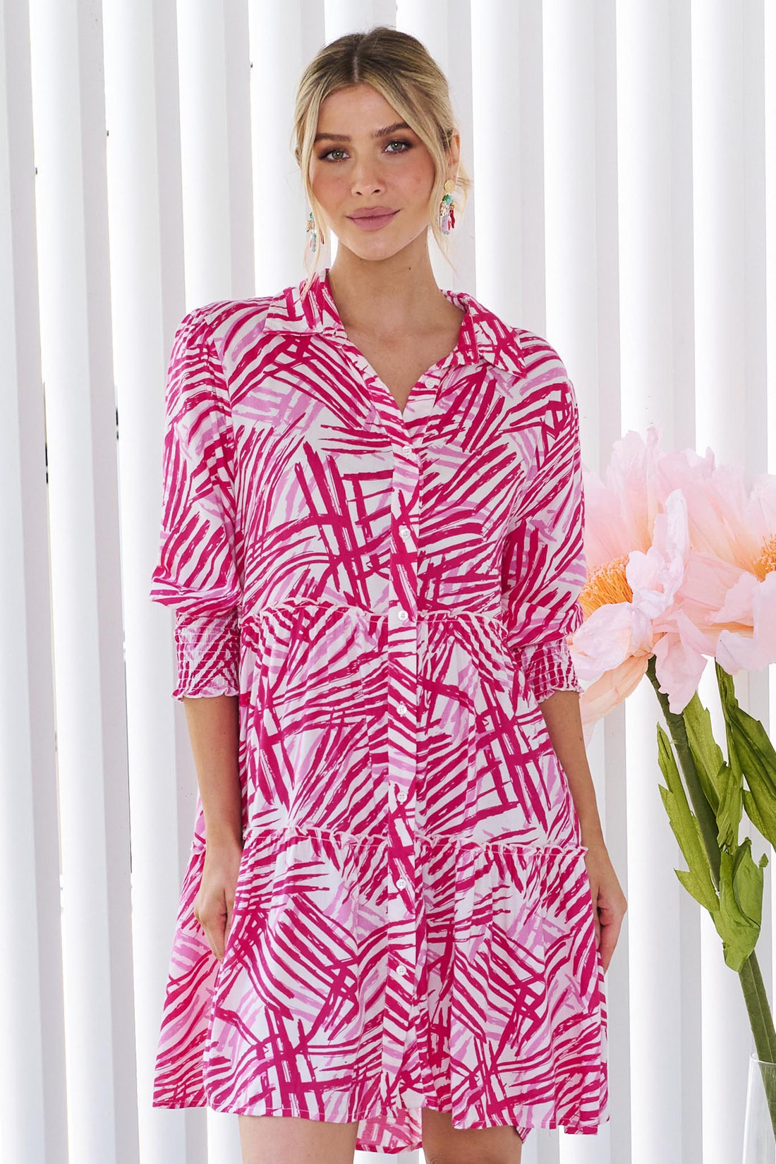 A woman wearing the minnie + MAXXIE Gemma pink and white abstract patterned mini dress with three-quarter sleeves, standing against a white panelled wall.