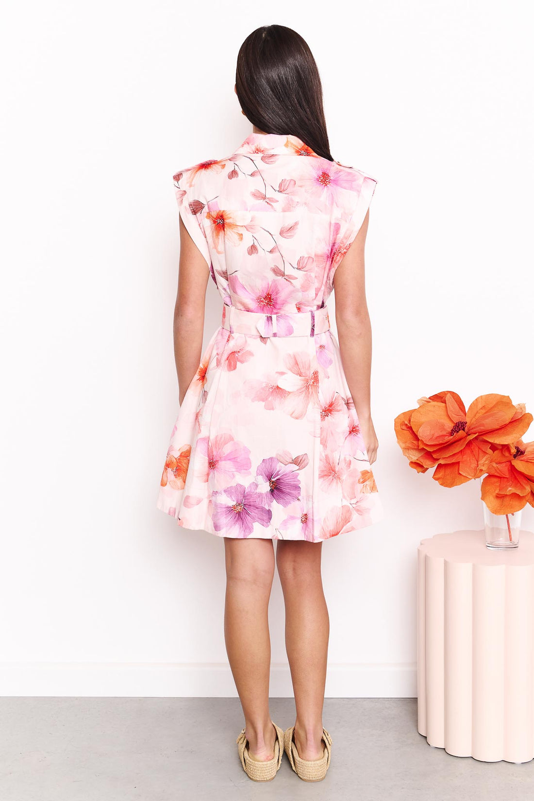 Back view of a woman wearing the minnie + MAXXIE Gracie sleeveless cotton dress with a pink and orange floral print, standing against a plain white background.