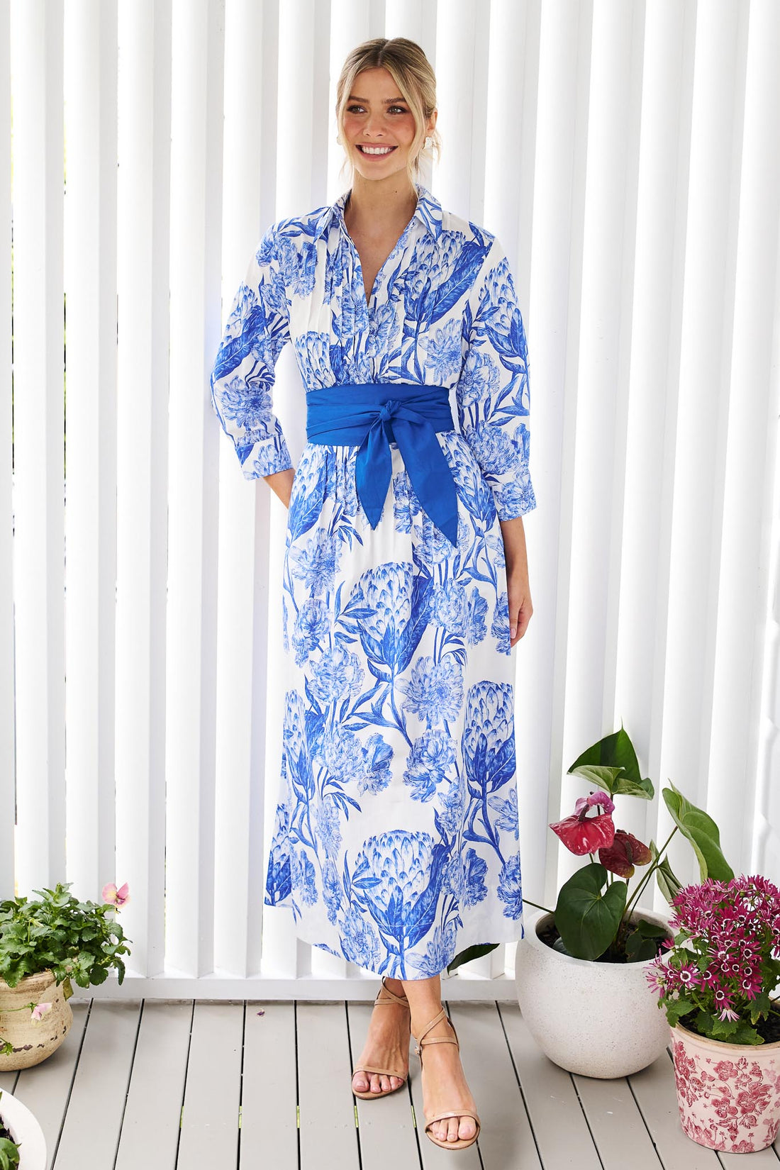 A woman wearing the minnie + MAXXIE Helena cotton midi dress with a blue floral print, pintuck pleats and a matching blue sash belt, standing in front of a white slatted wall with potted plants.