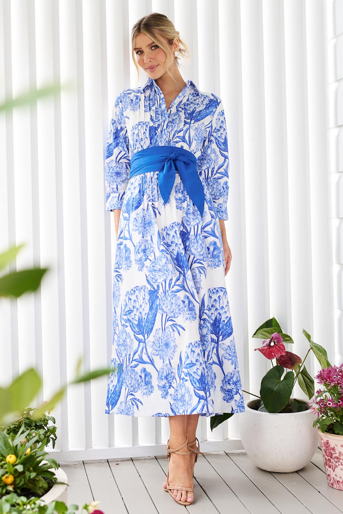 A woman wearing the minnie + MAXXIE Helena cotton midi dress with a blue floral print, pintuck pleats and a matching blue sash belt, standing in front of a white slatted wall with potted plants.
