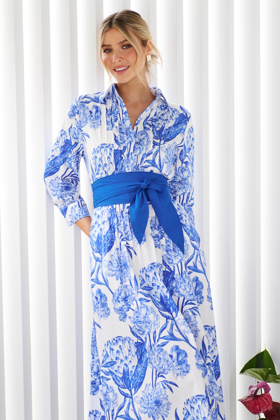 A woman wearing the minnie + MAXXIE Helena cotton midi dress with a blue floral print, pintuck pleats and a matching blue sash waist tie, standing in front of a white slatted wall.