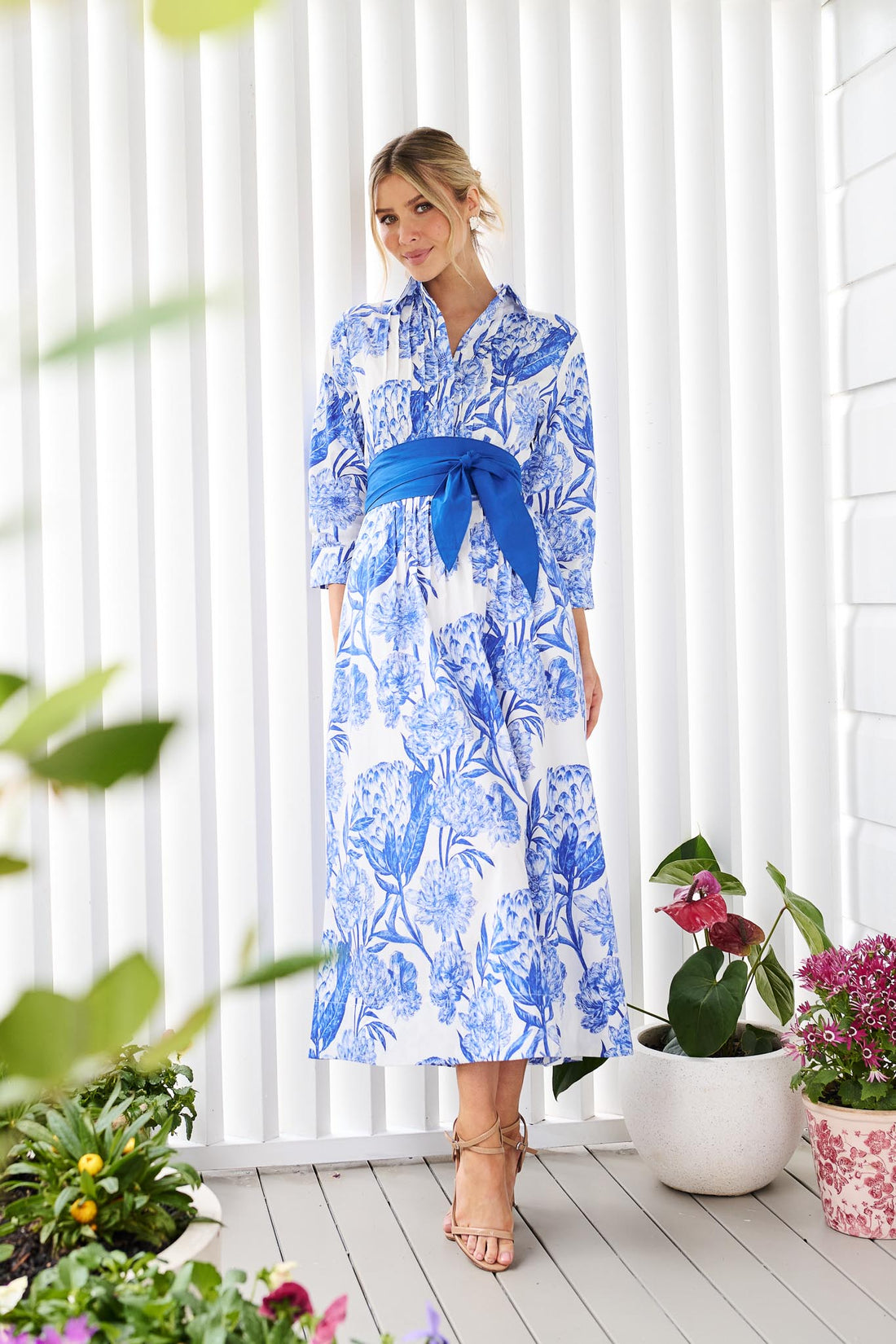 A woman wearing the minnie + MAXXIE Helena cotton midi dress with a blue floral print, pintuck pleats and a matching blue sash belt, standing on a white wooden deck in front of a white slatted wall with potted plants.