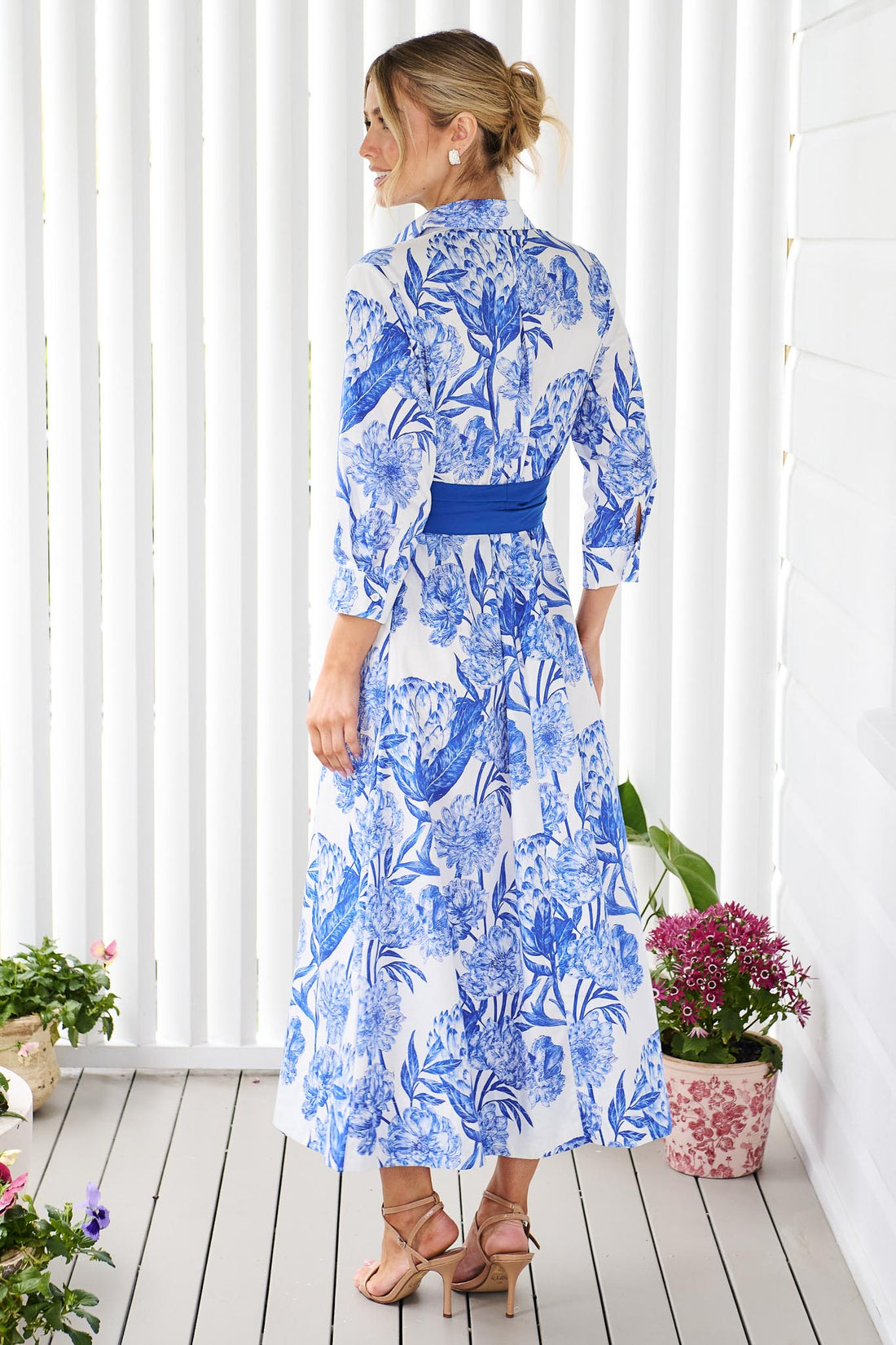 Back view of a woman wearing the minnie + MAXXIE Helena cotton midi dress with a blue floral print, pintuck pleats and a matching blue sash belt, standing on a white wooden deck in front of a white slatted wall with potted plants.