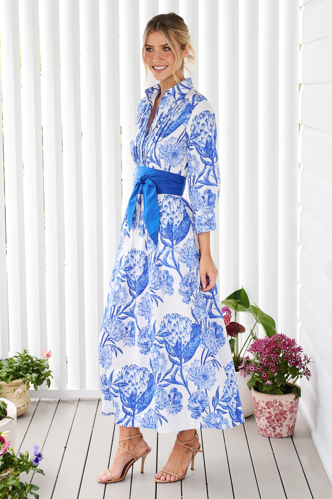 A woman wearing the minnie + MAXXIE Helena cotton midi dress with a blue floral print, pintuck pleats and a matching blue sash belt, standing on a white wooden deck in front of a white slatted wall with potted plants.