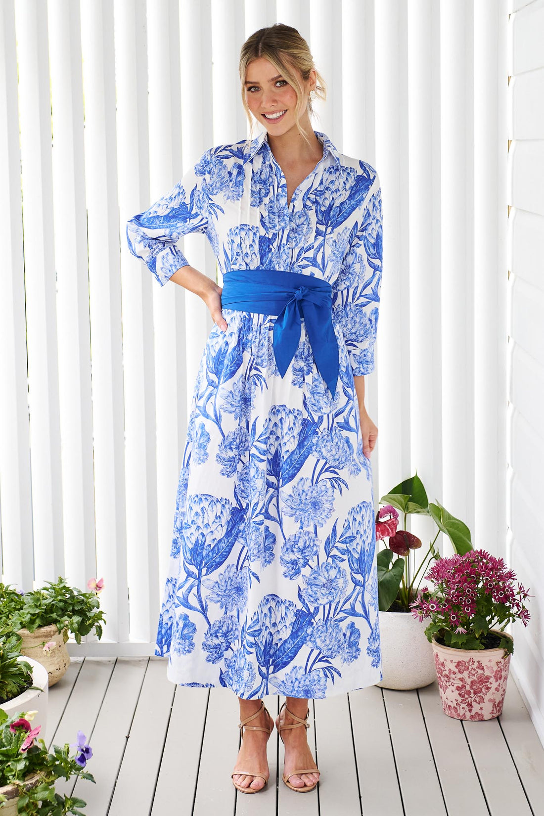A woman wearing the minnie + MAXXIE Helena cotton midi dress with a blue floral print, pintuck pleats and a matching blue sash belt, standing on a white wooden deck in front of a white slatted wall with potted plants.