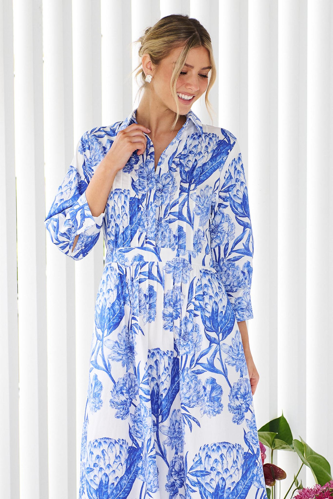 A woman wearing the minnie + MAXXIE Helena cotton midi dress with a blue floral print, standing in front of a white slatted wall with potted plants.
