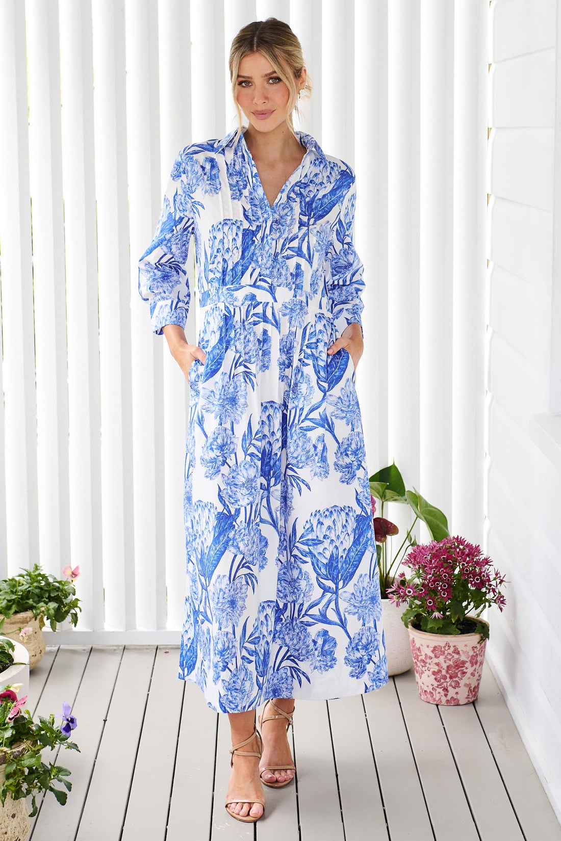 A woman wearing the minnie + MAXXIE Helena cotton midi dress with a blue floral print, standing on a white wooden deck in front of a white slatted wall with potted plants.