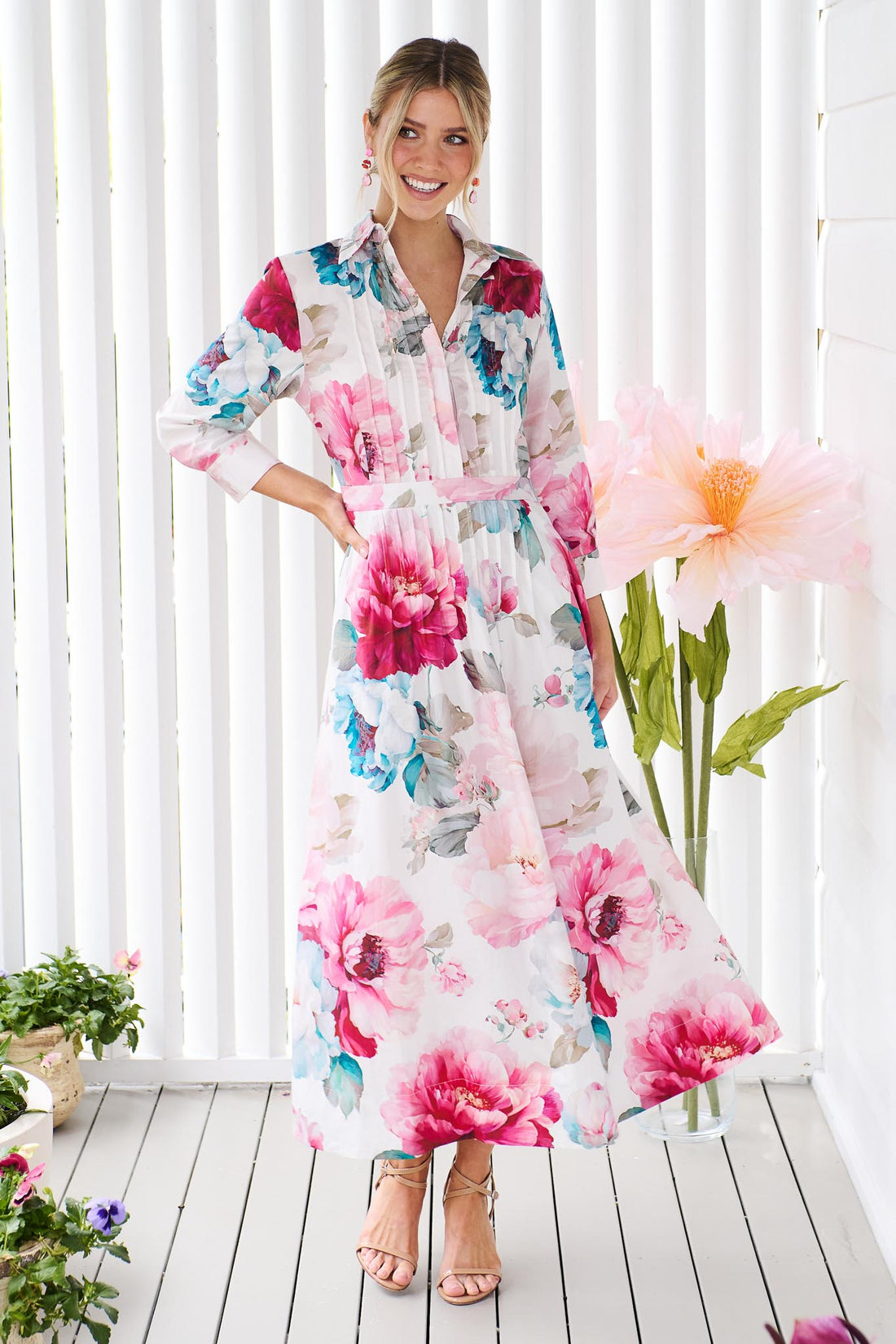 A woman wearing the minnie + MAXXIE Helena cotton midi dress with a pink floral print and pintuck pleats, standing in front of a white slatted wall with potted plants.