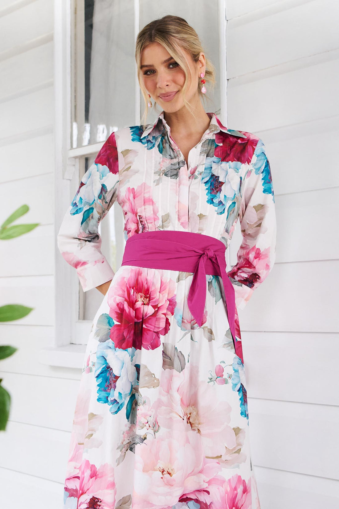 Clos up of a woman wearing the minnie + MAXXIE Helena cotton midi dress with a pink floral print, pintuck pleats and a matching magenta pink sash waist tie, standing in front of a white window.
