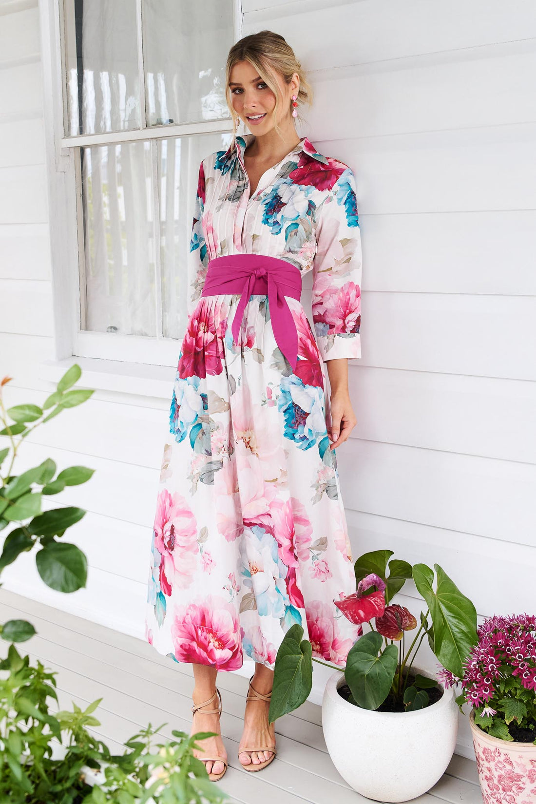 A woman wearing the minnie + MAXXIE Helena cotton midi dress with a pink floral print, pintuck pleats and a matching magenta pink sash waist tie, standing in front of a white slatted wall with potted plants.