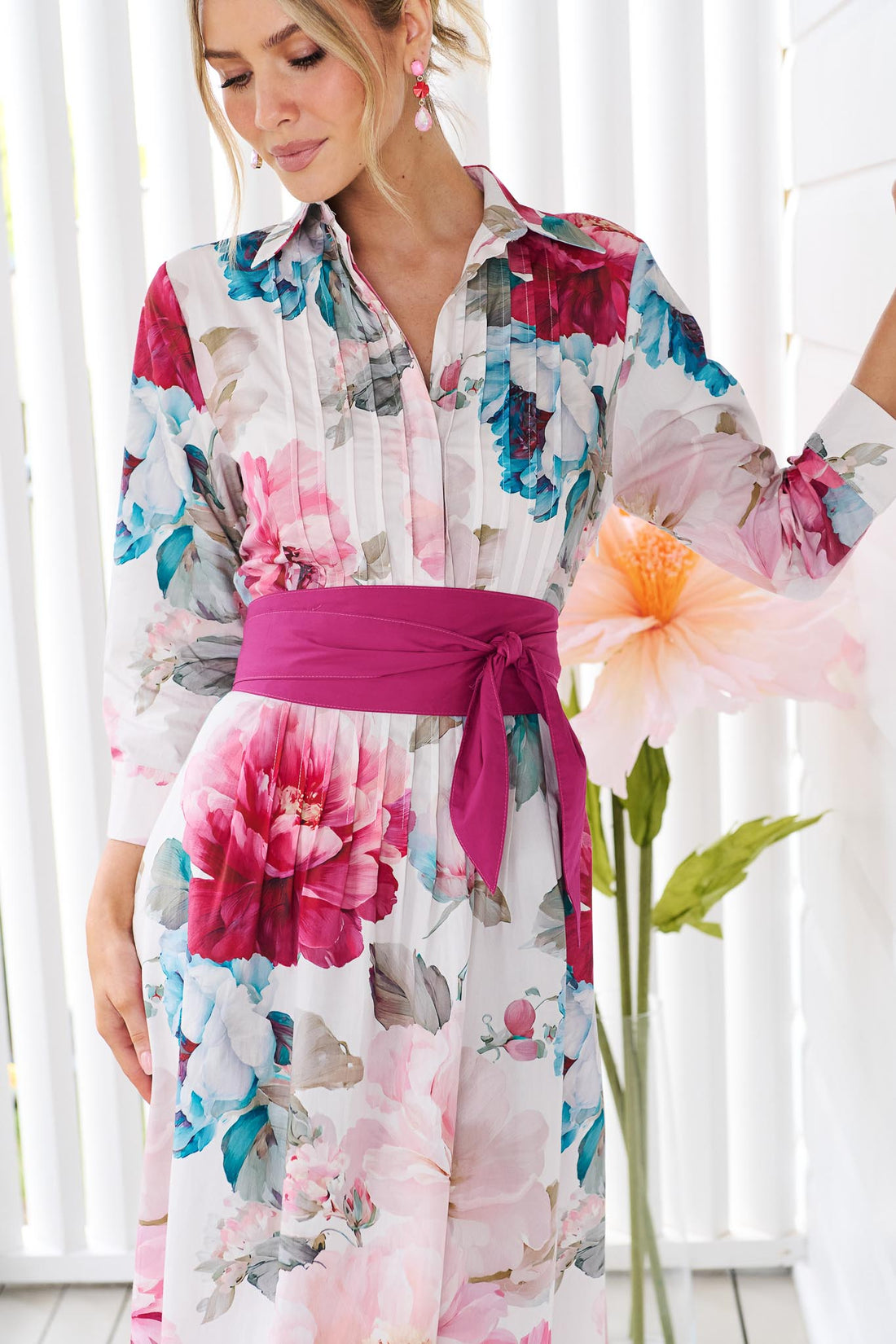 Detail of a woman wearing the minnie + MAXXIE Helena cotton midi dress with a pink floral print, pintuck pleats and a matching magenta pink sash waist tie, standing in front of a white slatted wall.