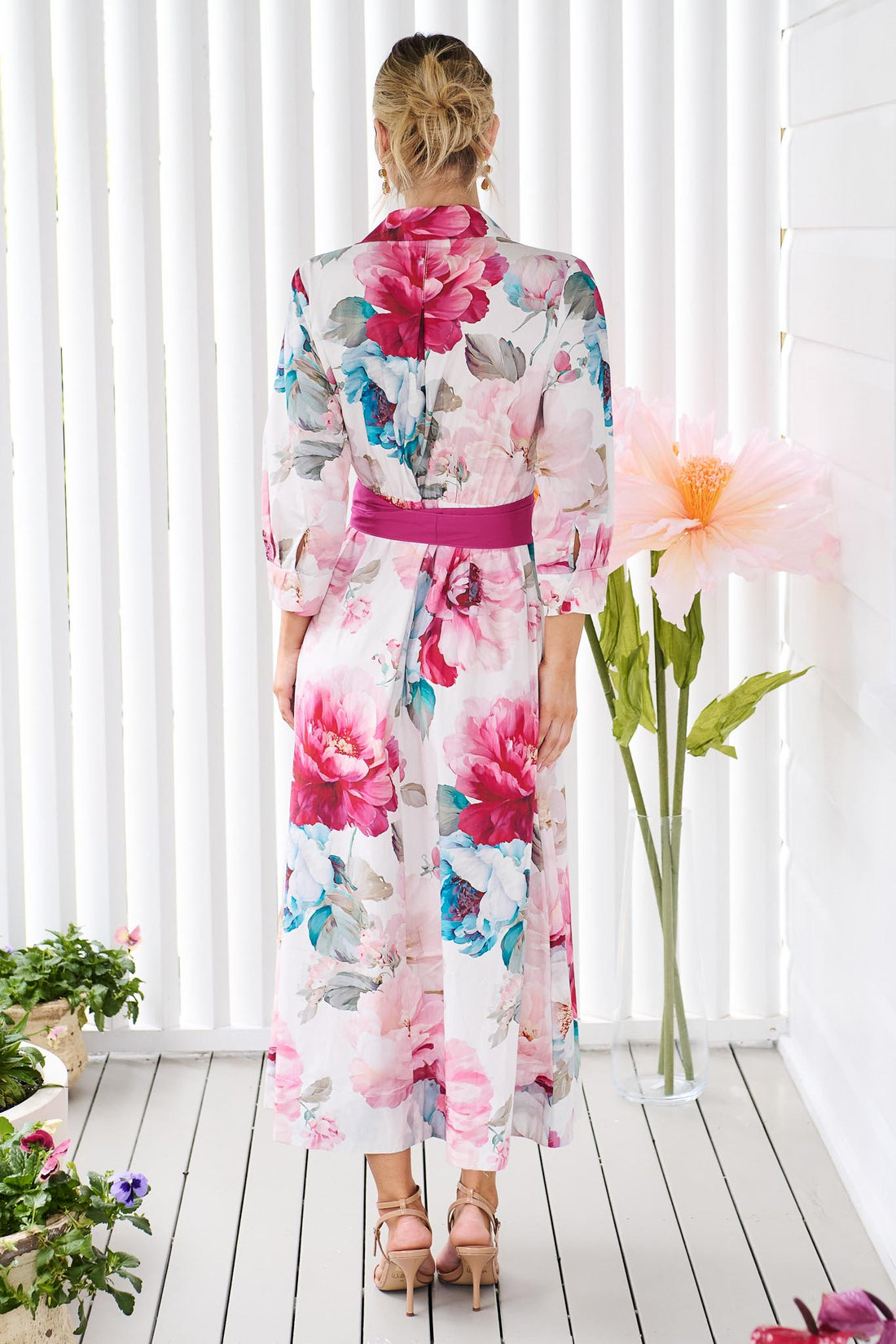 Back view of a woman wearing the minnie + MAXXIE Helena cotton midi dress with a pink floral print, pintuck pleats and a matching magenta pink sash waist tie, standing in front of a white slatted wall with potted plants.