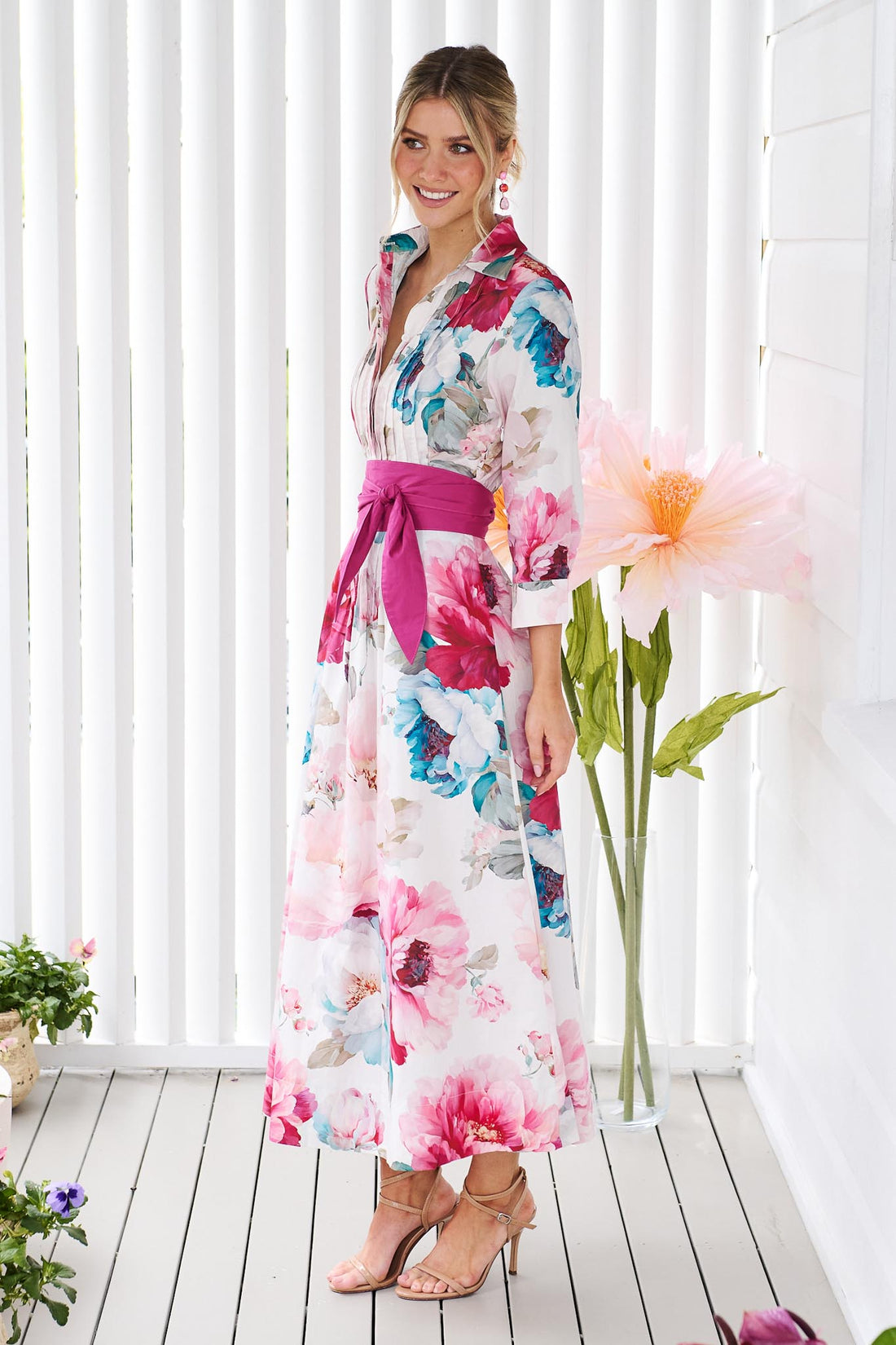 Side view of a woman wearing the minnie + MAXXIE Helena cotton midi dress with a pink floral print, pintuck pleats and a matching magenta pink sash waist tie, standing in front of a white slatted wall with potted plants.