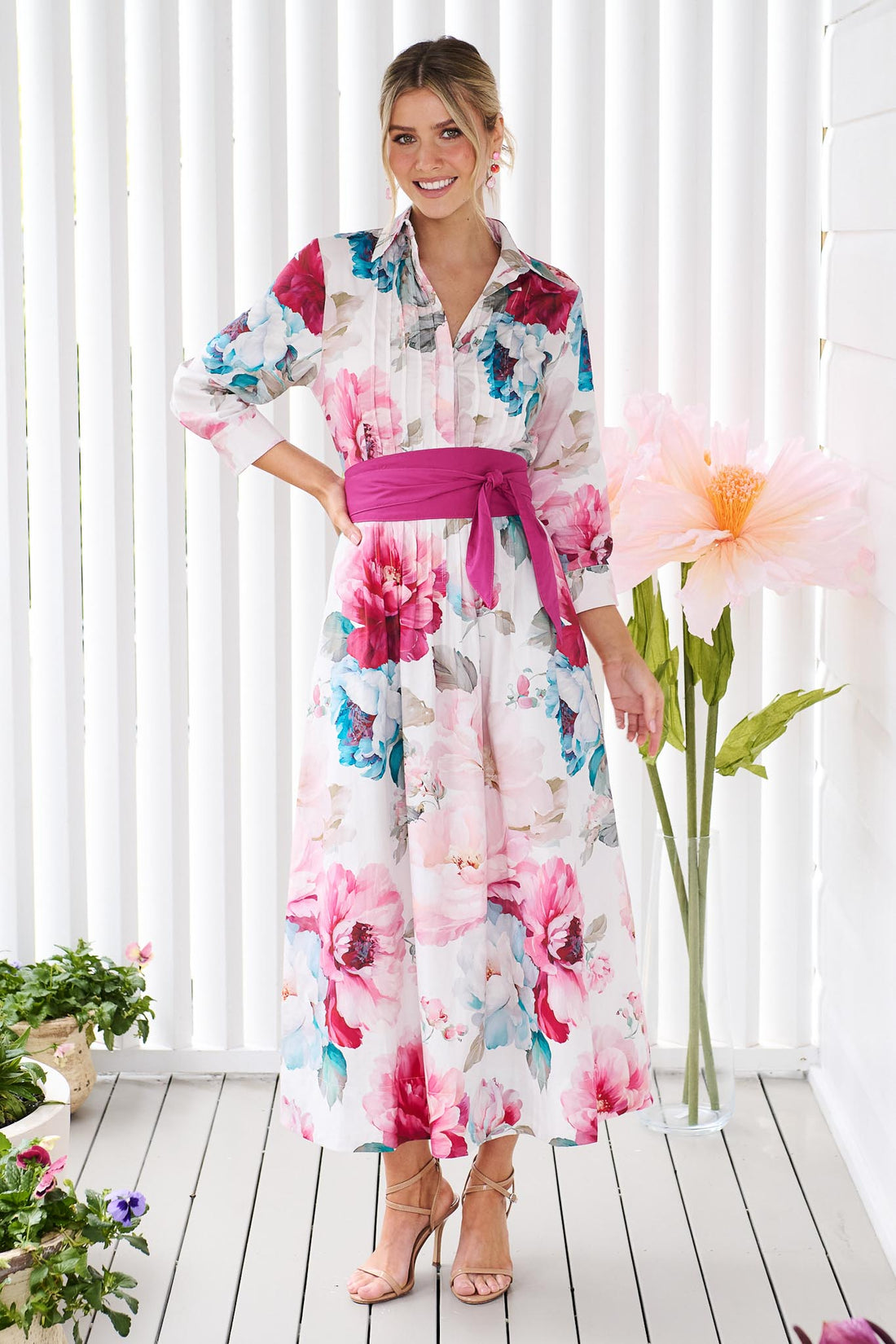 A woman wearing the minnie + MAXXIE Helena cotton midi dress with a pink floral print, pintuck pleats and a matching magenta pink sash waist tie, standing in front of a white slatted wall with potted plants.