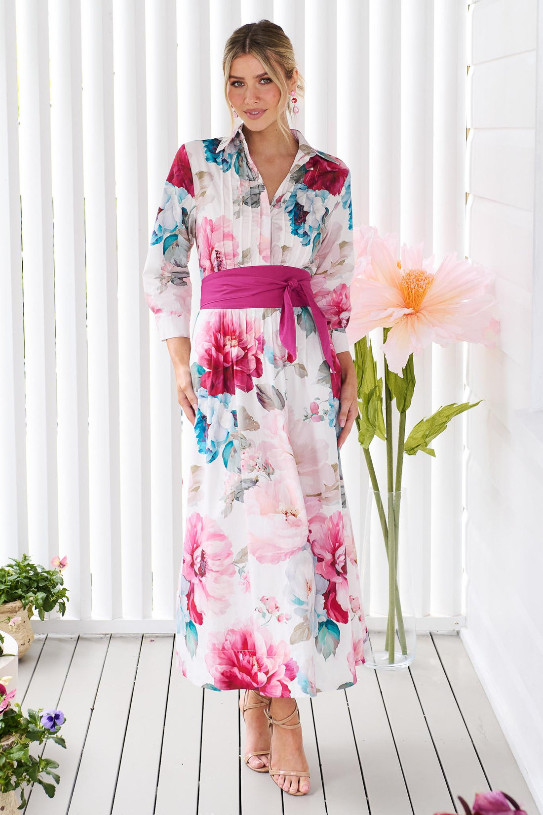 A woman wearing the minnie + MAXXIE Helena cotton midi dress with a pink floral print, pintuck pleats and a matching magenta pink sash waist tie, standing in front of a white slatted wall with potted plants.