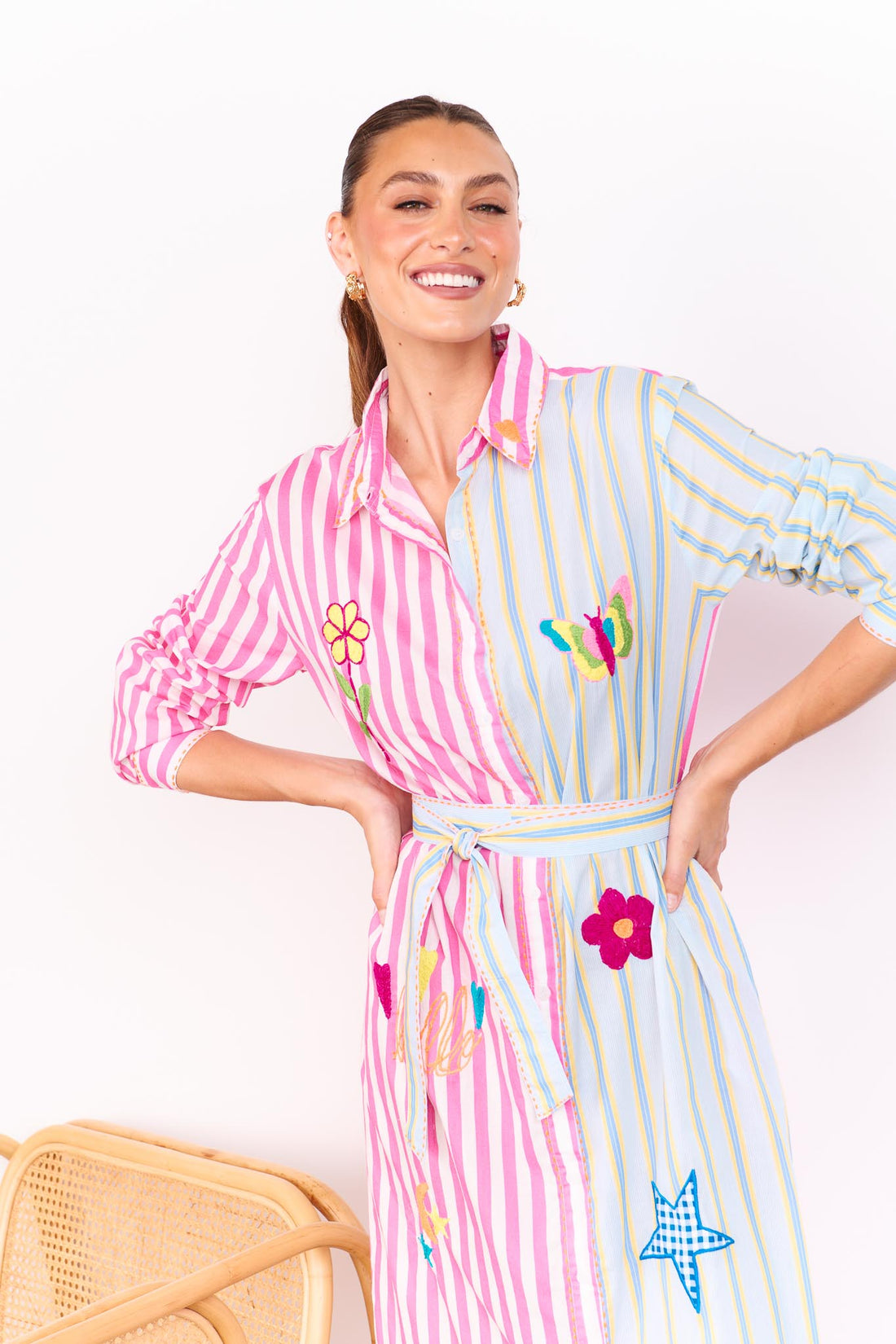 Woman wearing the minnie + MAXXIE Hello Sunshine Cotton midi shirt dress with long sleeves and a matching waist tie. One side is pink stripes and the other side blue pinstripes. She is standing in front of a white wall.