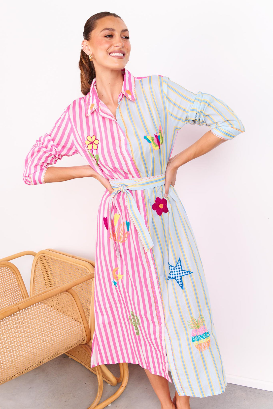 Woman wearing the minnie + MAXXIE Hello Sunshine Cotton midi shirt dress with long sleeves and a matching waist tie. One side is pink stripes and the other side blue pinstripes. She is standing in front of a white wall.