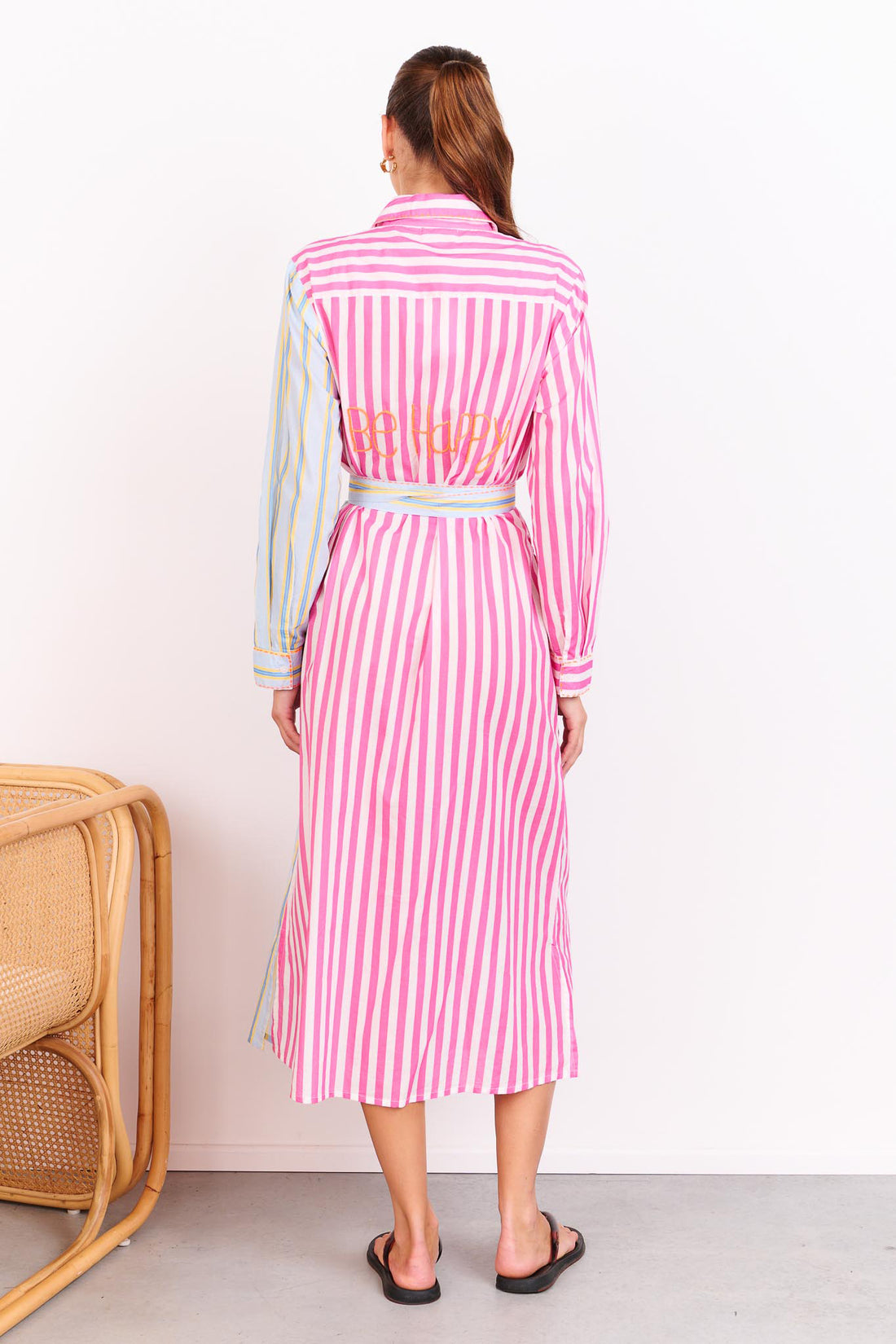 Back view of a woman wearing the minnie + MAXXIE Hello Sunshine Cotton midi shirt dress with long sleeves and a matching waist tie. She is standing in front of a white wall beside a cane chair.