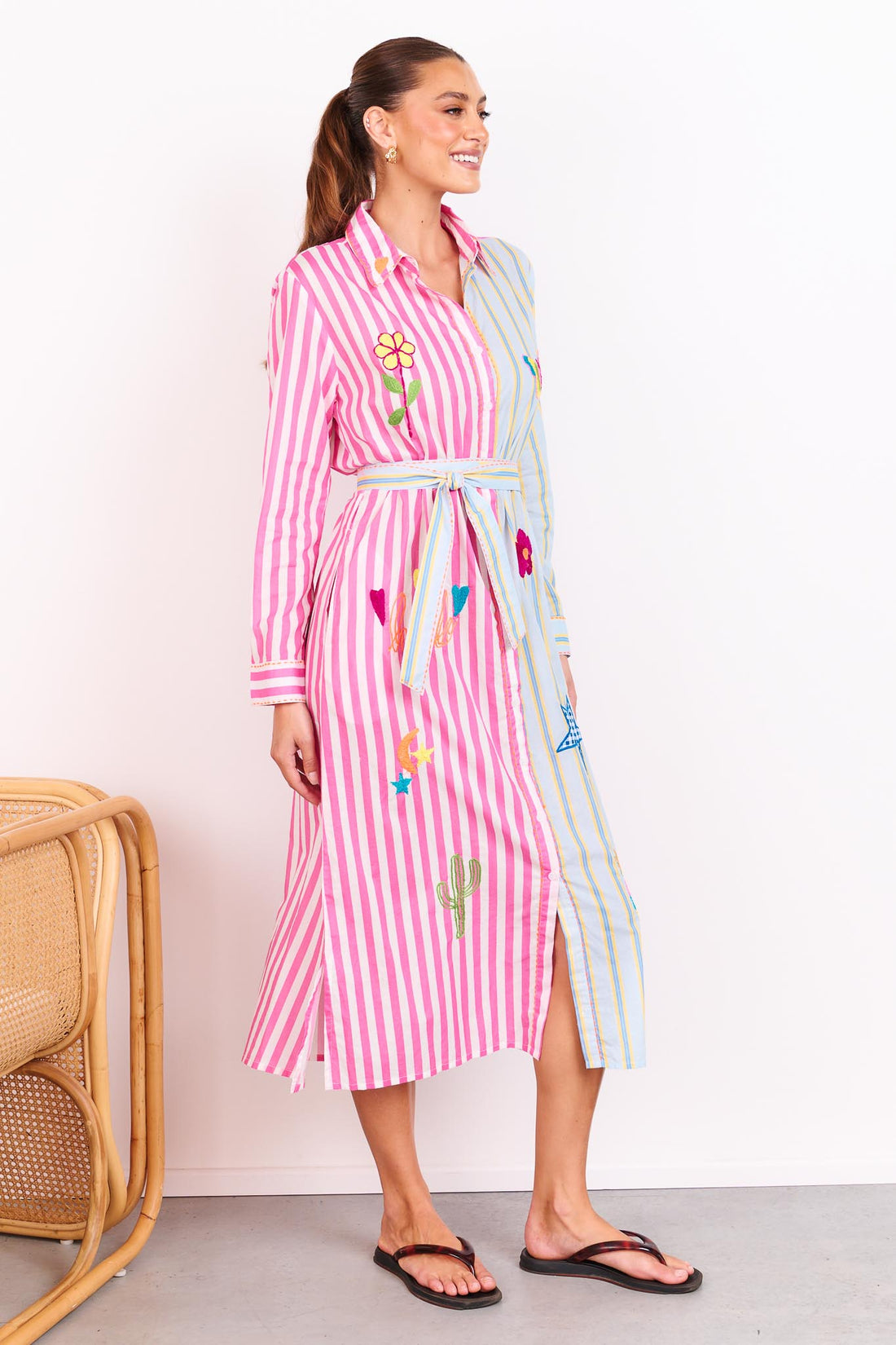Woman wearing the minnie + MAXXIE Hello Sunshine Cotton midi shirt dress with long sleeves and a matching waist tie. One side is pink stripes and the other side blue pinstripes. She is standing in front of a white wall.