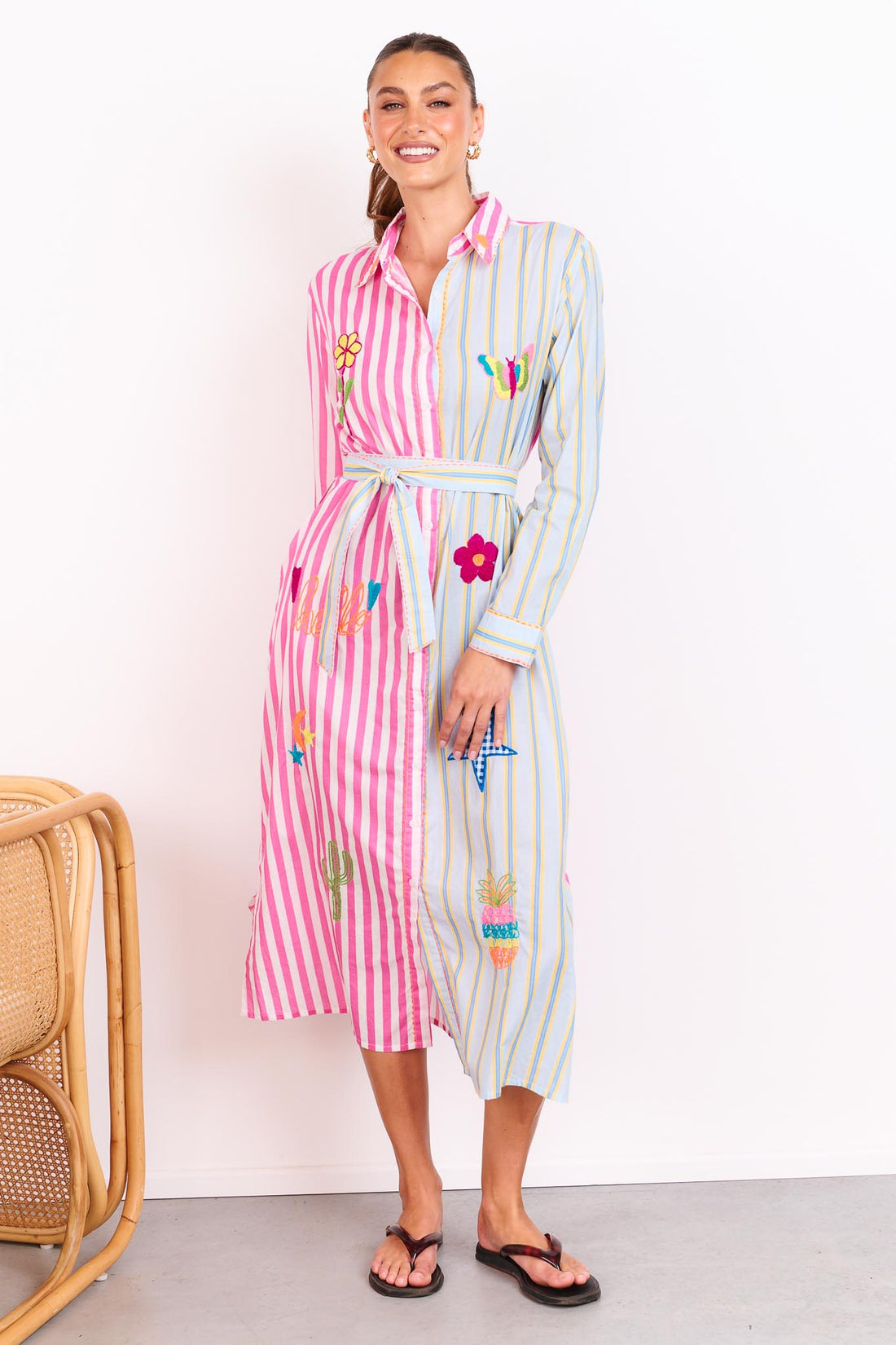 Woman wearing the minnie + MAXXIE Hello Sunshine Cotton midi shirt dress with long sleeves and a matching waist tie. One side is pink stripes and the other side blue pinstripes. She is standing in front of a white wall.