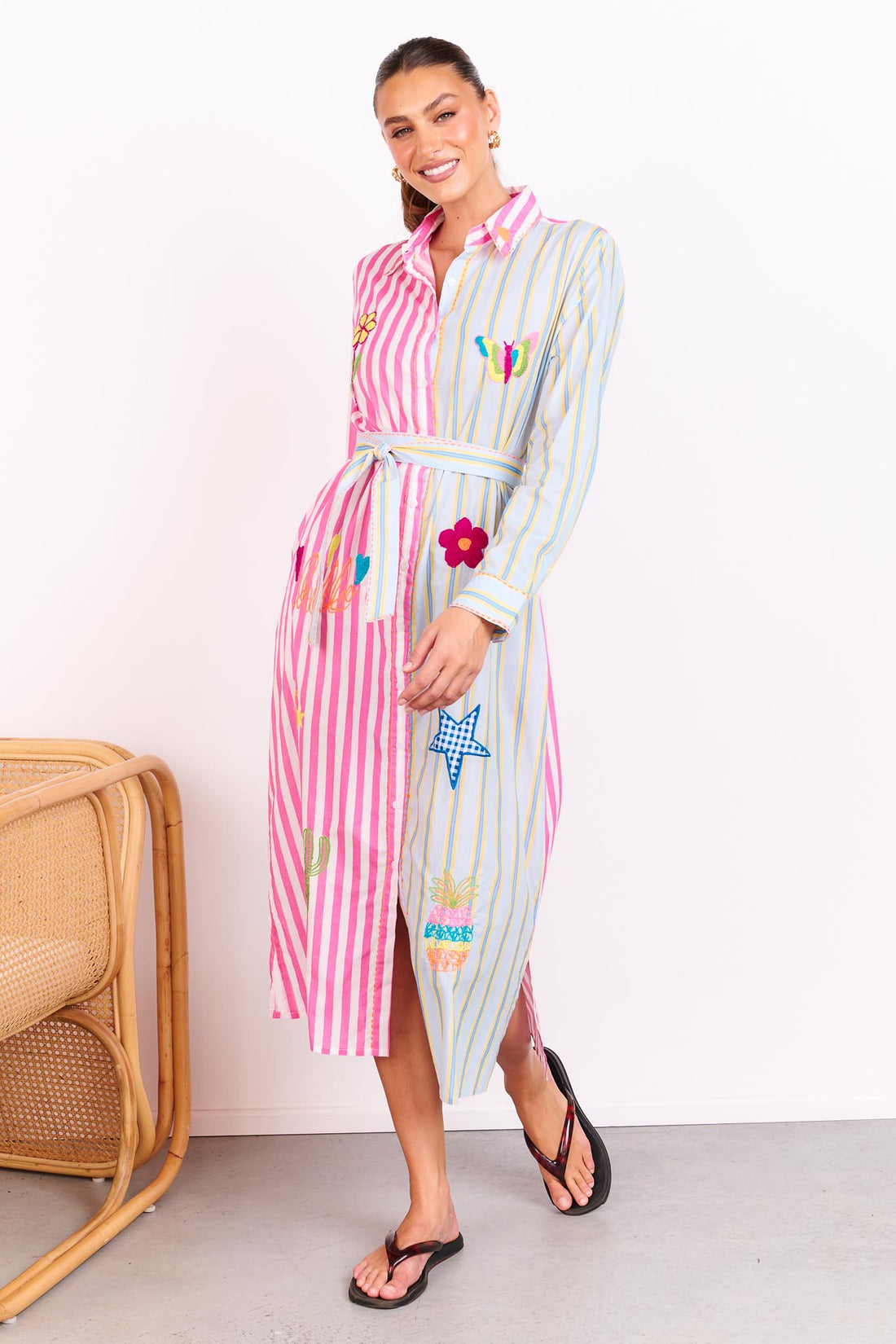 Woman wearing the minnie + MAXXIE Hello Sunshine Cotton midi shirt dress with long sleeves and a matching waist tie. One side is pink stripes and the other side blue pinstripes. She is standing in front of a white wall.