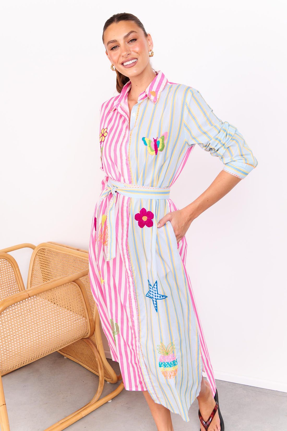 Woman wearing the minnie + MAXXIE Hello Sunshine Cotton midi shirt dress with long sleeves and a matching waist tie. One side is pink stripes and the other side blue pinstripes. She is standing in front of a white wall.