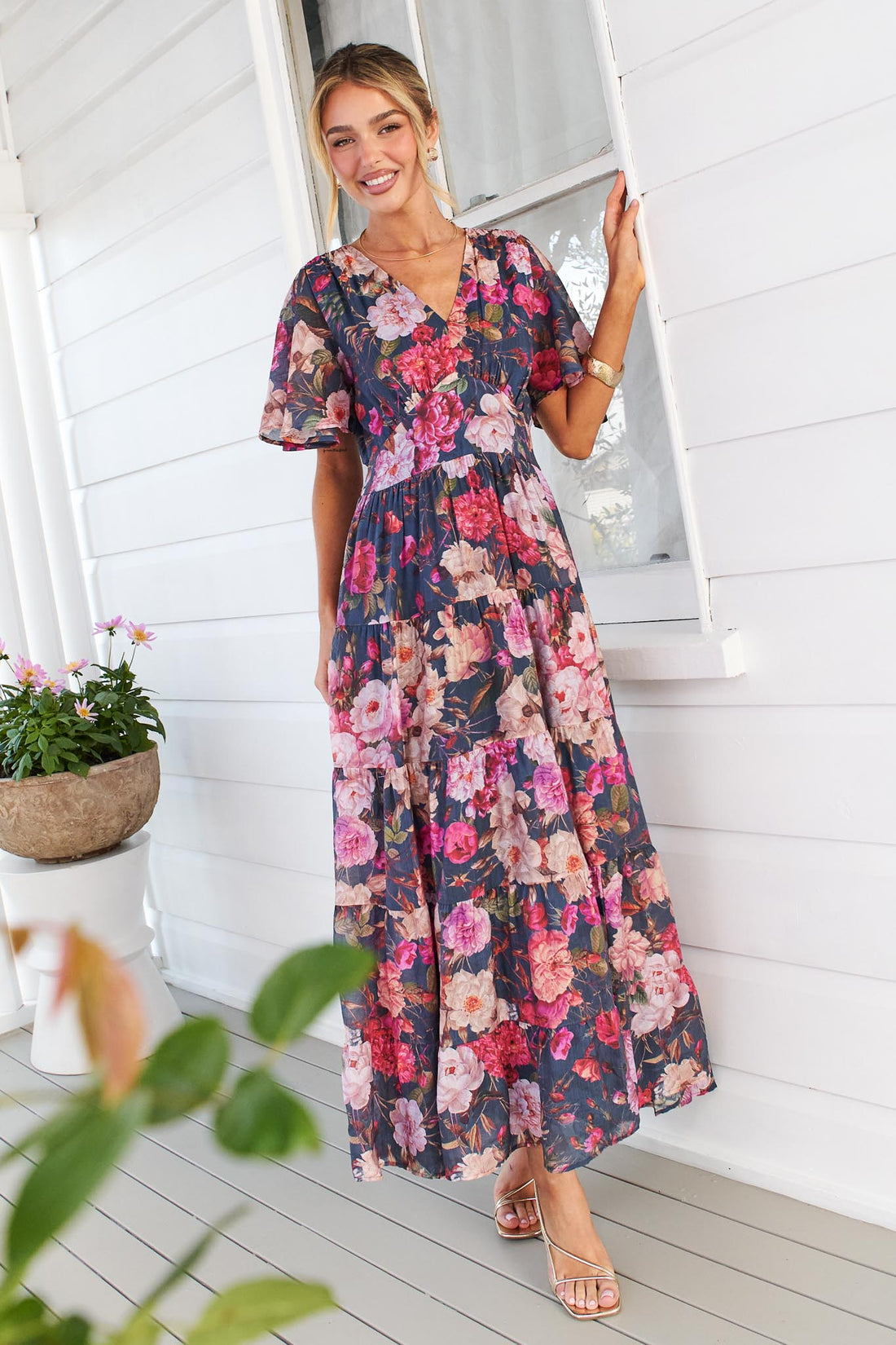 A woman models the short-sleeve, V-neck minnie + MAXXIE Hollie maxi dress with a rose print on a dark navy background, standing on a white porch next to a window.