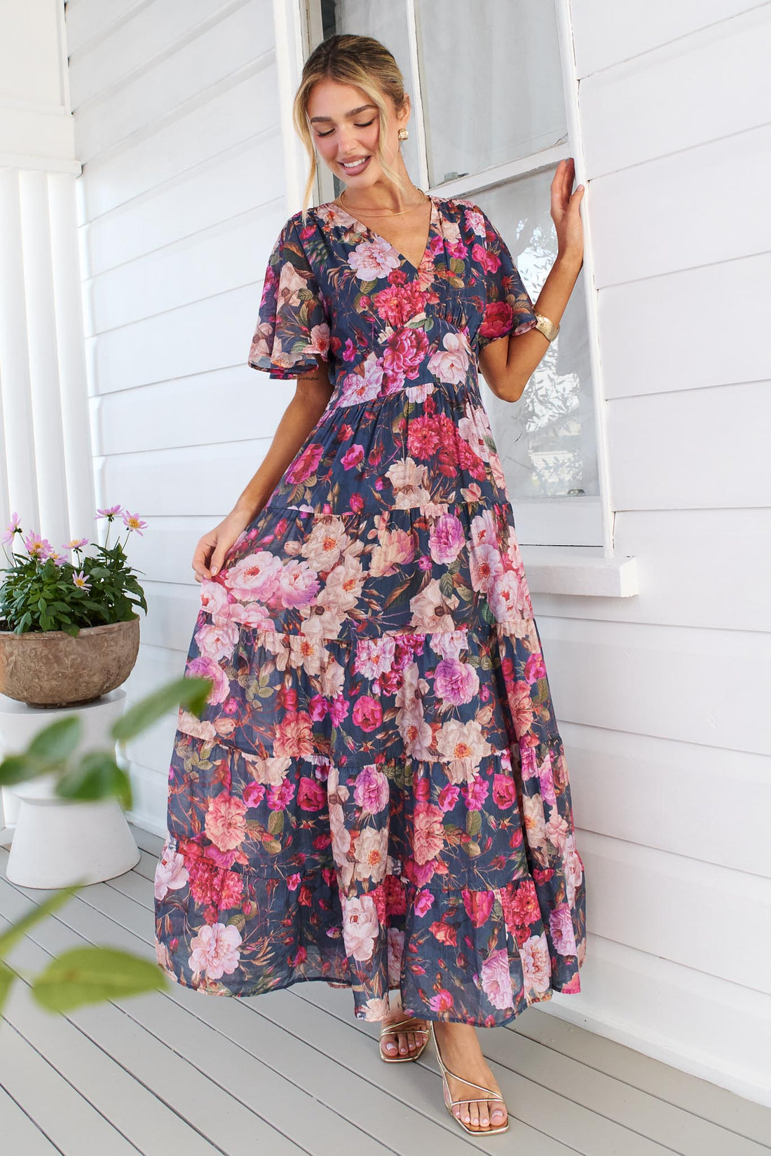 A woman models the short-sleeve, V-neck minnie + MAXXIE Hollie maxi dress with a rose print on a dark navy background, standing on a white porch next to a window.
