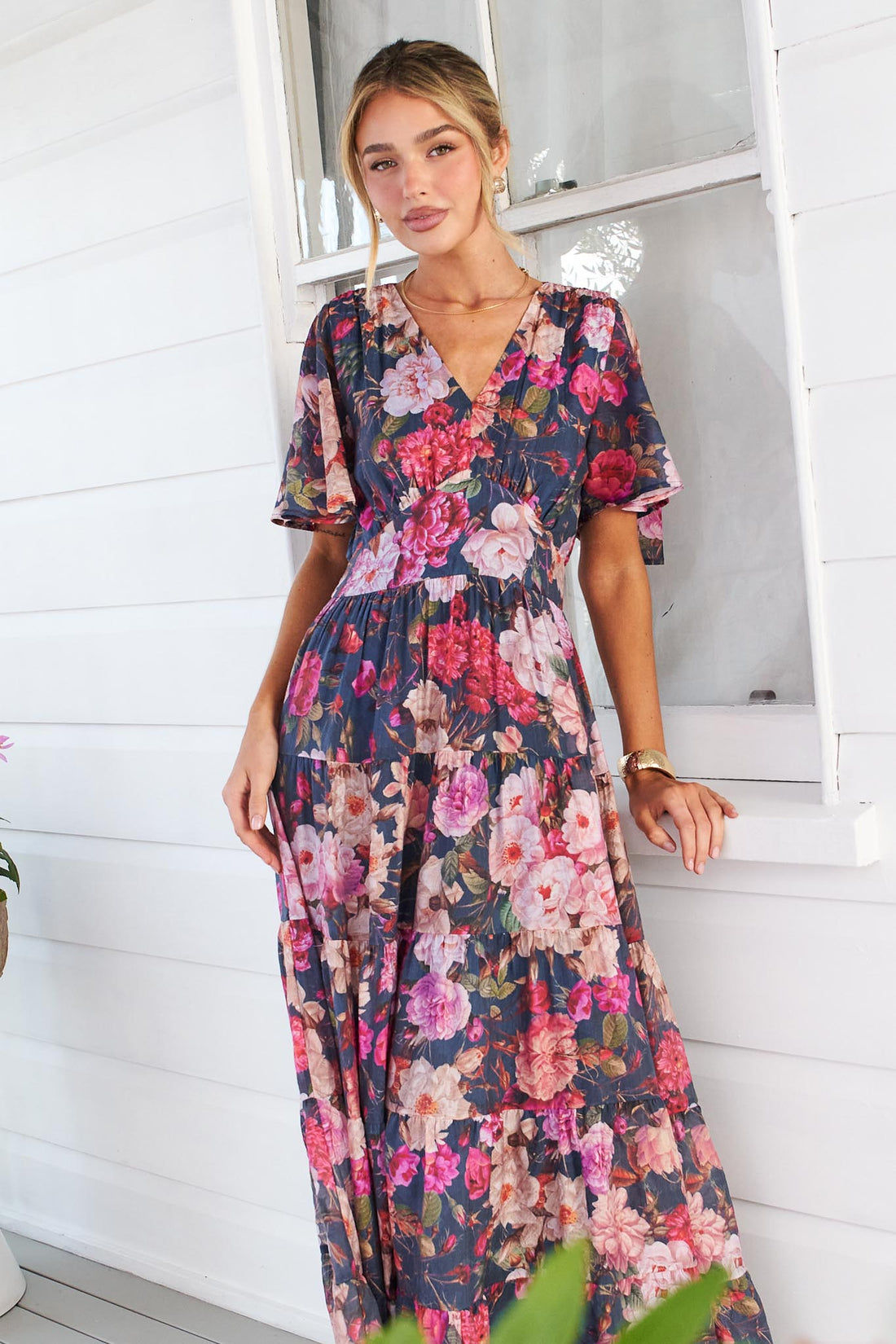 A woman models the short-sleeve, V-neck minnie + MAXXIE Hollie maxi dress with a rose print on a dark navy background, standing on a white porch next to a window.