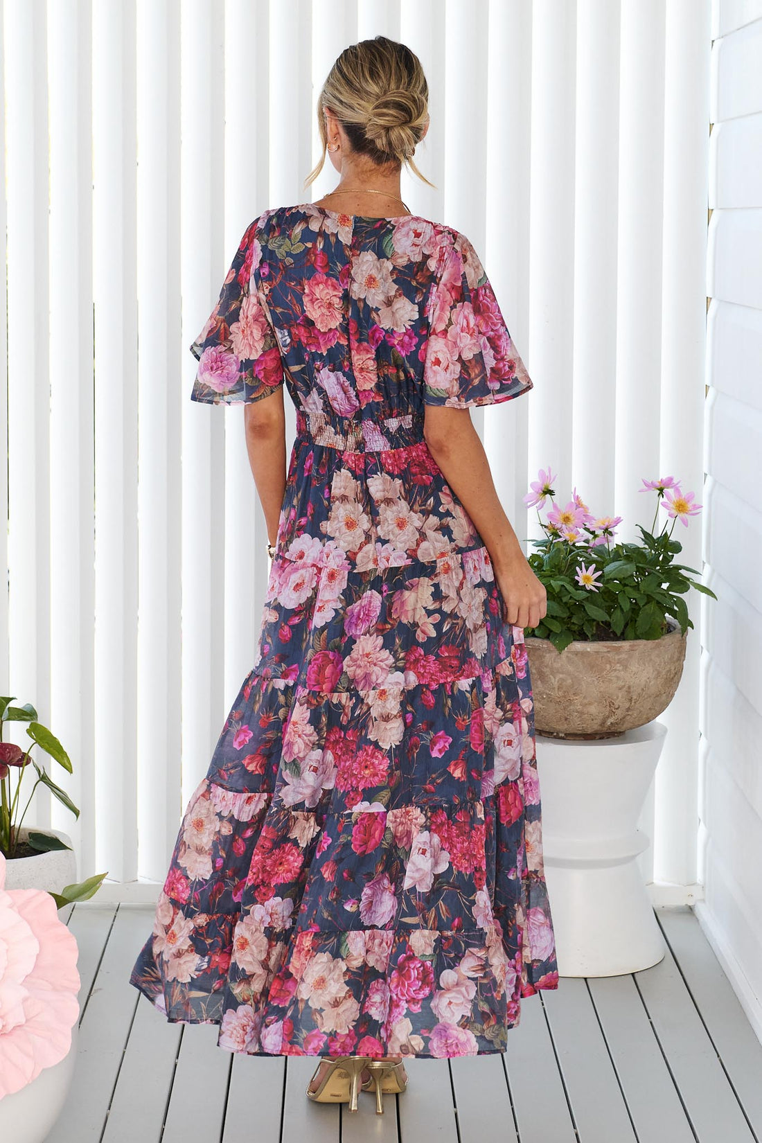 Back view of a woman modelling the short-sleeve, V-neck minnie + MAXXIE Hollie maxi dress with a rose print on a dark navy background, standing in front of a white panelled wall.