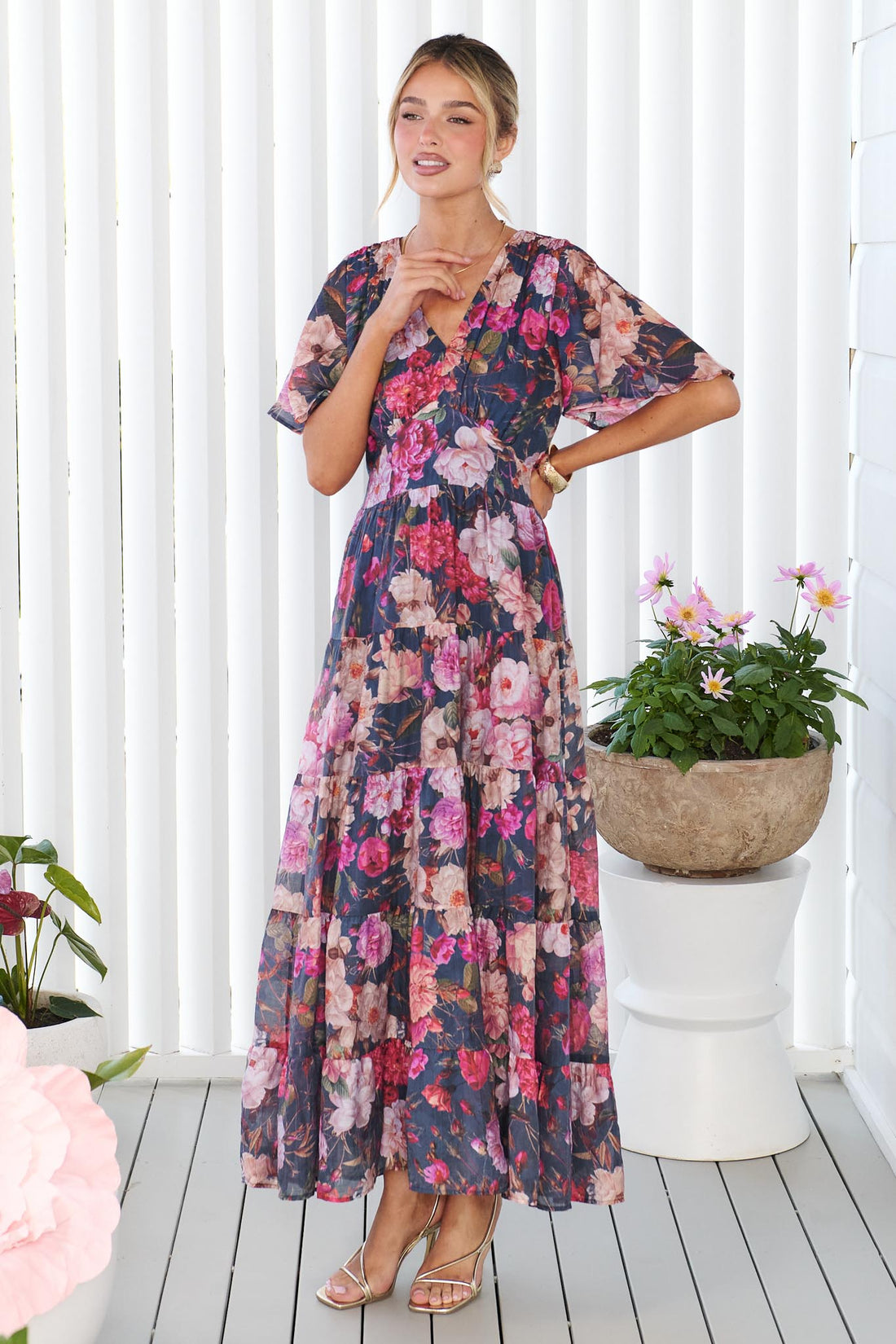 A woman models the short-sleeve, V-neck minnie + MAXXIE Hollie maxi dress with a rose print on a dark navy background, standing in front of a white panelled wall.
