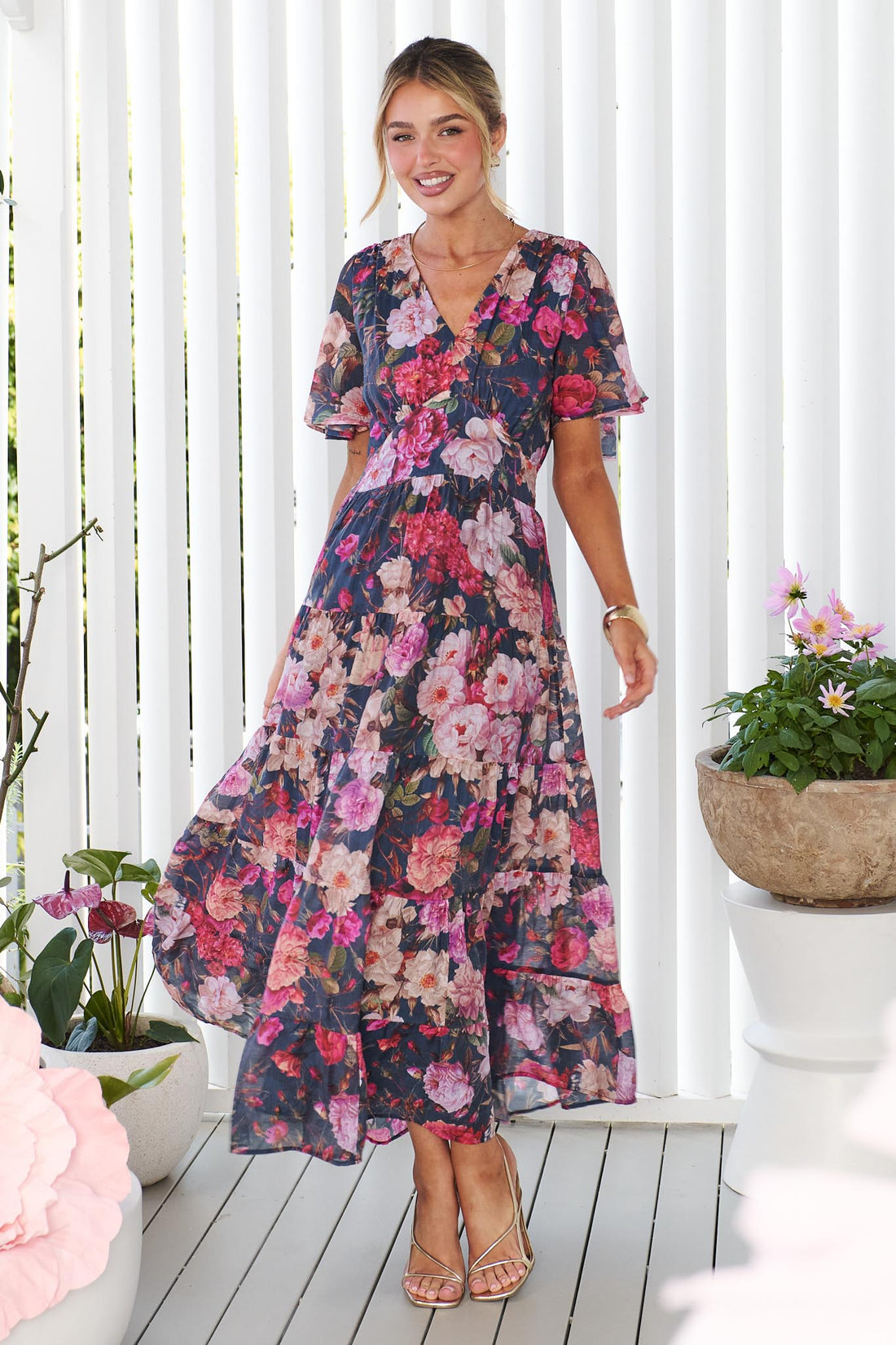 A woman models the short-sleeve, V-neck minnie + MAXXIE Hollie maxi dress with a rose print on a dark navy background, standing in front of a white panelled wall.