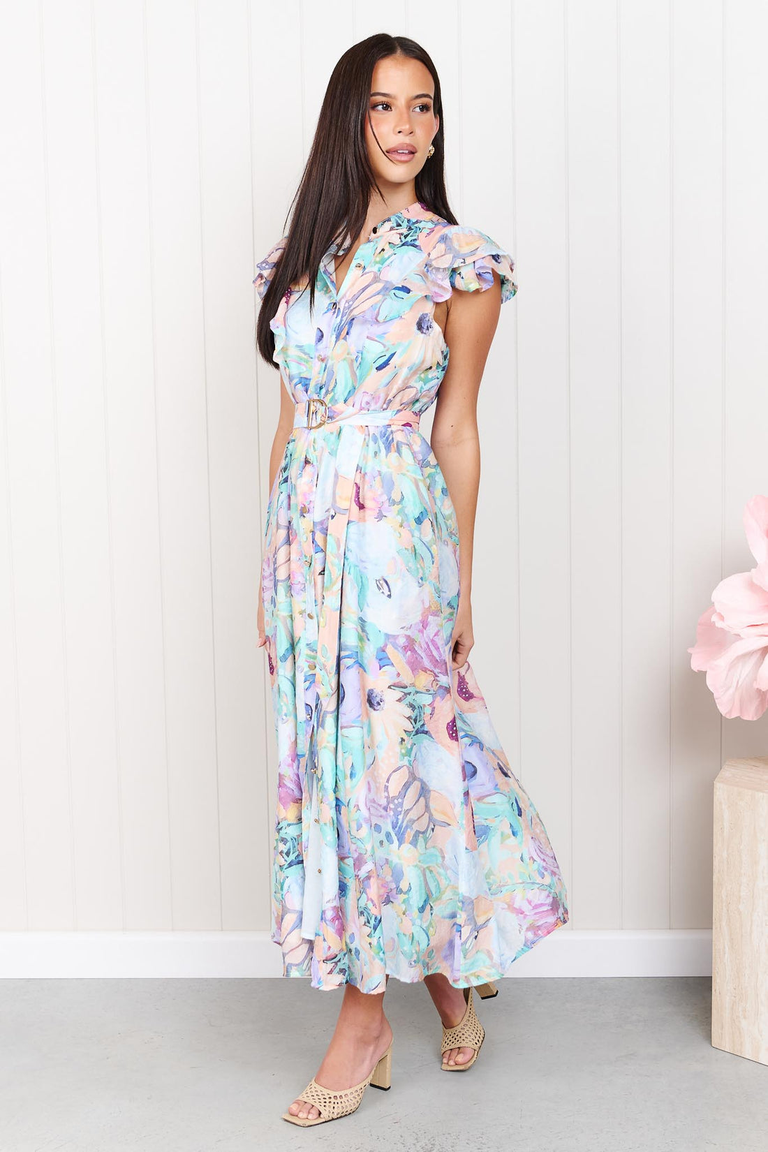 A woman wearing the minnie + MAXXIE Jardin Midi Dress in a pastel watercolour floral print with ruffled cap sleeves and a matching belt, standing against a white panelled wall.