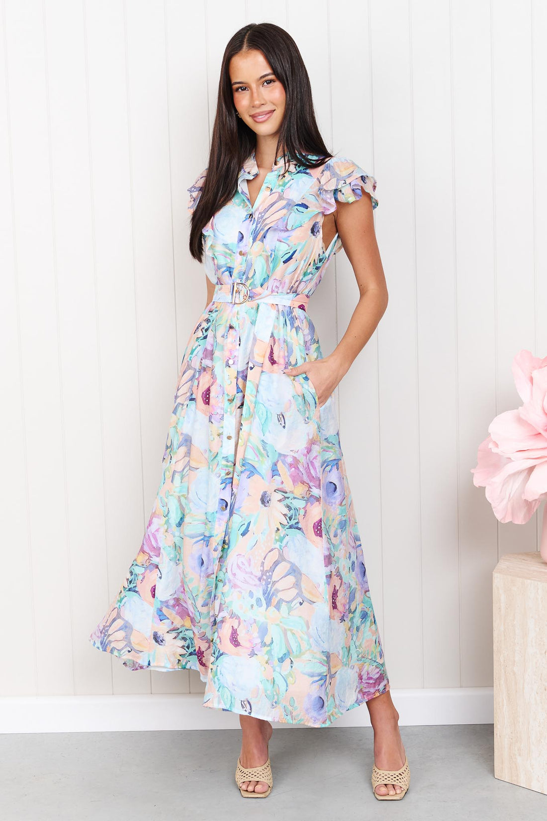 A woman wearing the minnie + MAXXIE Jardin Midi Dress in a pastel watercolour floral print with ruffled cap sleeves and a matching belt, standing against a white panelled wall.