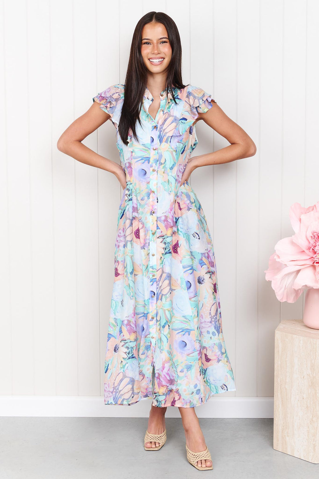A woman wearing the minnie + MAXXIE Jardin Midi Dress in a pastel watercolour floral print with ruffled cap sleeves, standing against a white panelled wall.