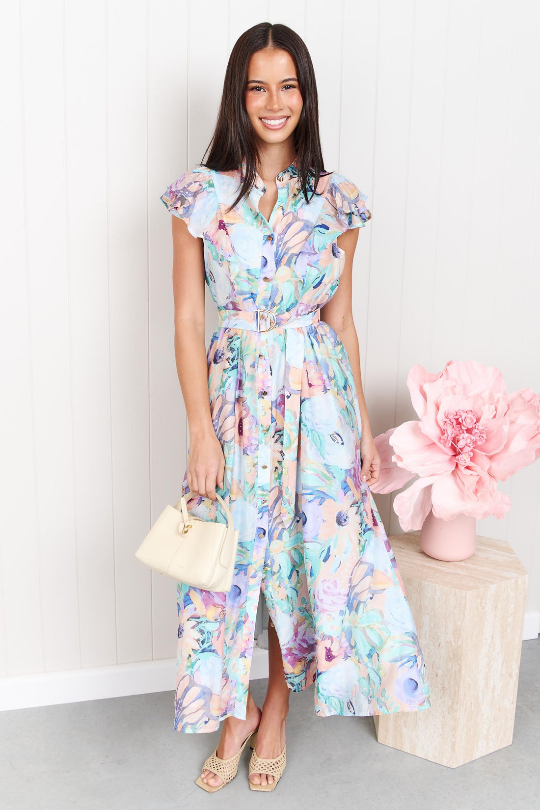 A woman wearing the minnie + MAXXIE Jardin Midi Dress in a pastel watercolour floral print with ruffled cap sleeves and a matching belt, standing against a white panelled wall.