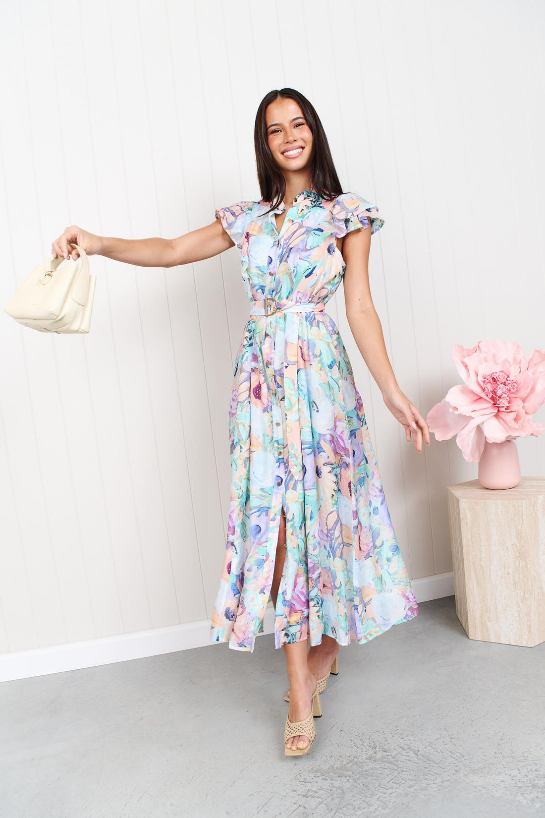 A woman wearing the minnie + MAXXIE Jardin Midi Dress in a pastel watercolour floral print with ruffled cap sleeves and a matching belt, standing against a white panelled wall.