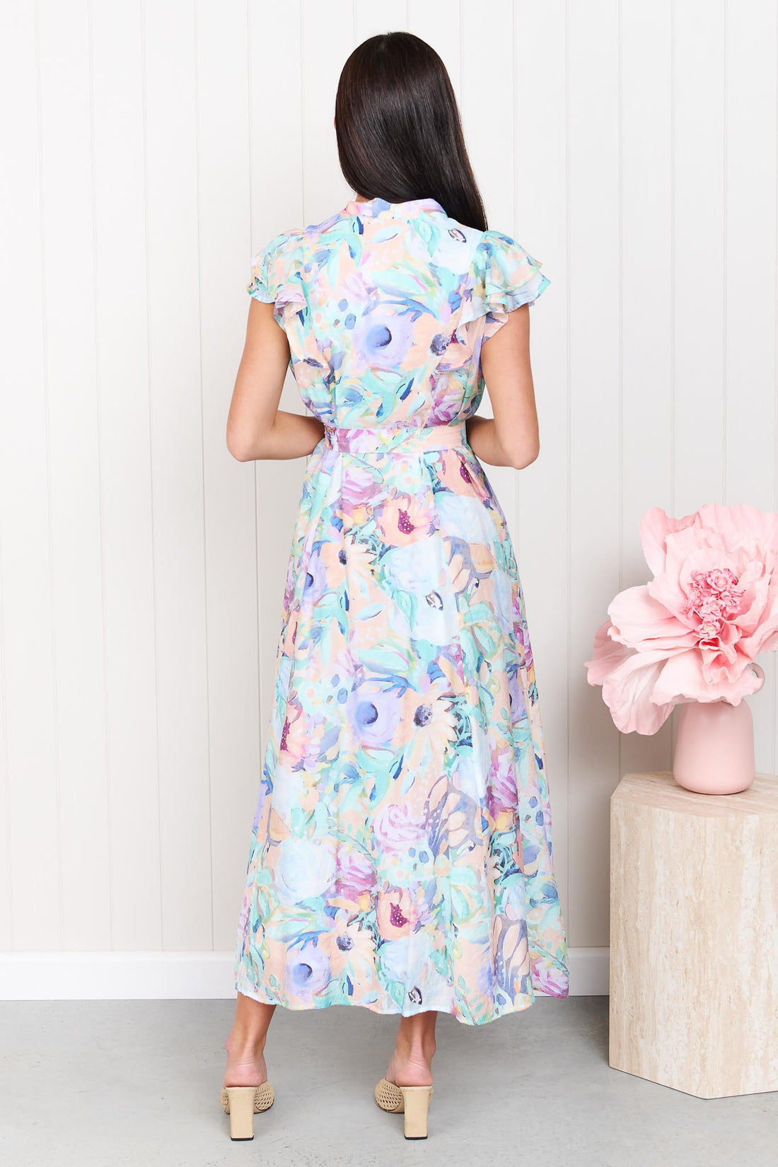 Back view of a woman wearing the minnie + MAXXIE Jardin Midi Dress in a pastel watercolour floral print with ruffled cap sleeves and a matching belt, standing against a white panelled wall.