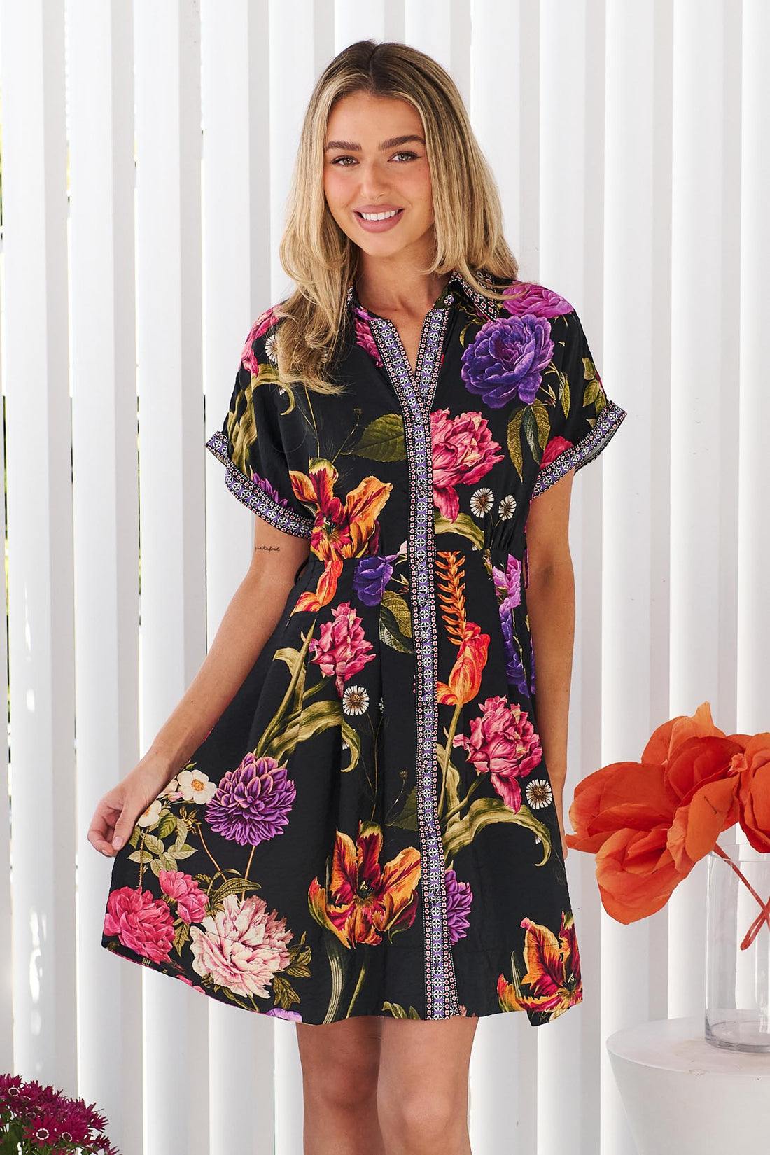 A woman wearing the minnie + MAXXIE Jemima dress with a vibrant multicolour floral print on a black background, set against a white panelled wall.