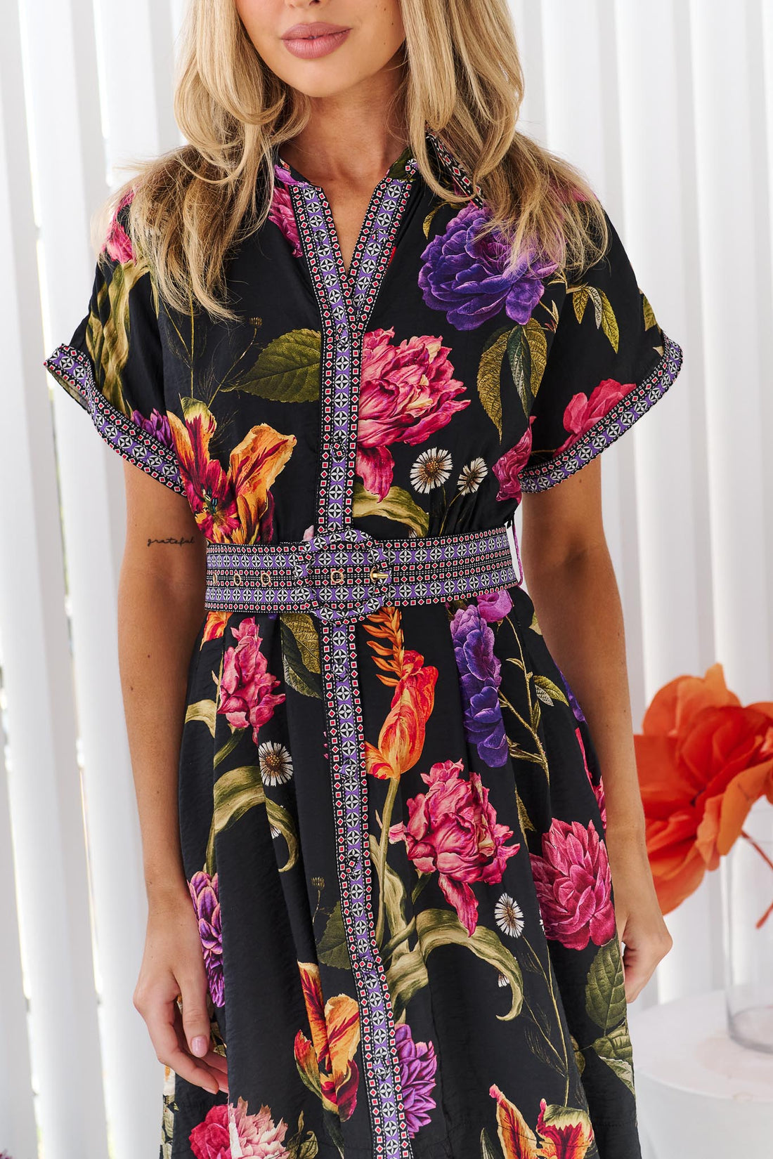 Detail of a woman wearing the minnie + MAXXIE Jemima dress with a vibrant multicolour floral print on a black background and a patterned waist tie, set against a white panelled wall.