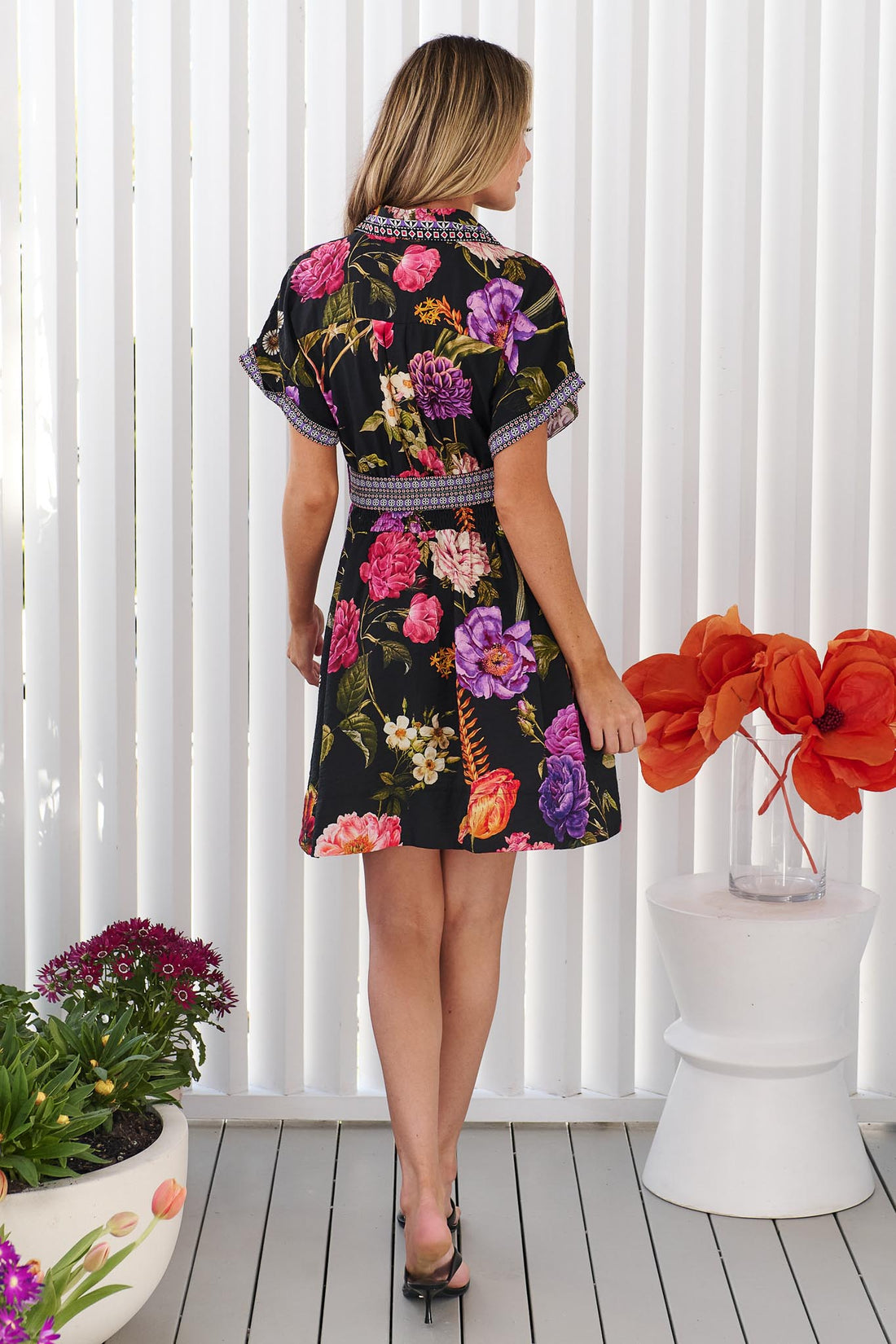 Back view of a woman wearing the minnie + MAXXIE Jemima dress with a vibrant multicolour floral print on a black background and a patterned waist tie, set against a white panelled wall.
