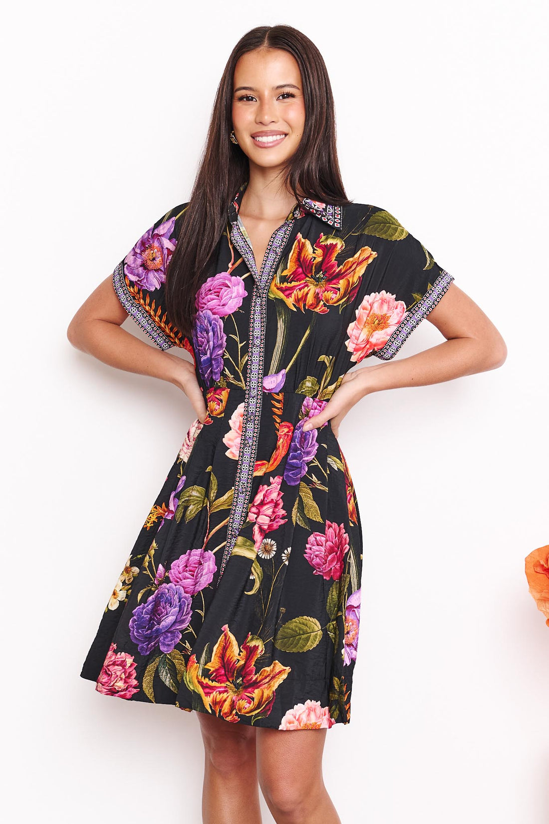 A woman wearing the minnie + MAXXIE Jemima dress with a vibrant multicolour floral print on a black background, set against a white wall.