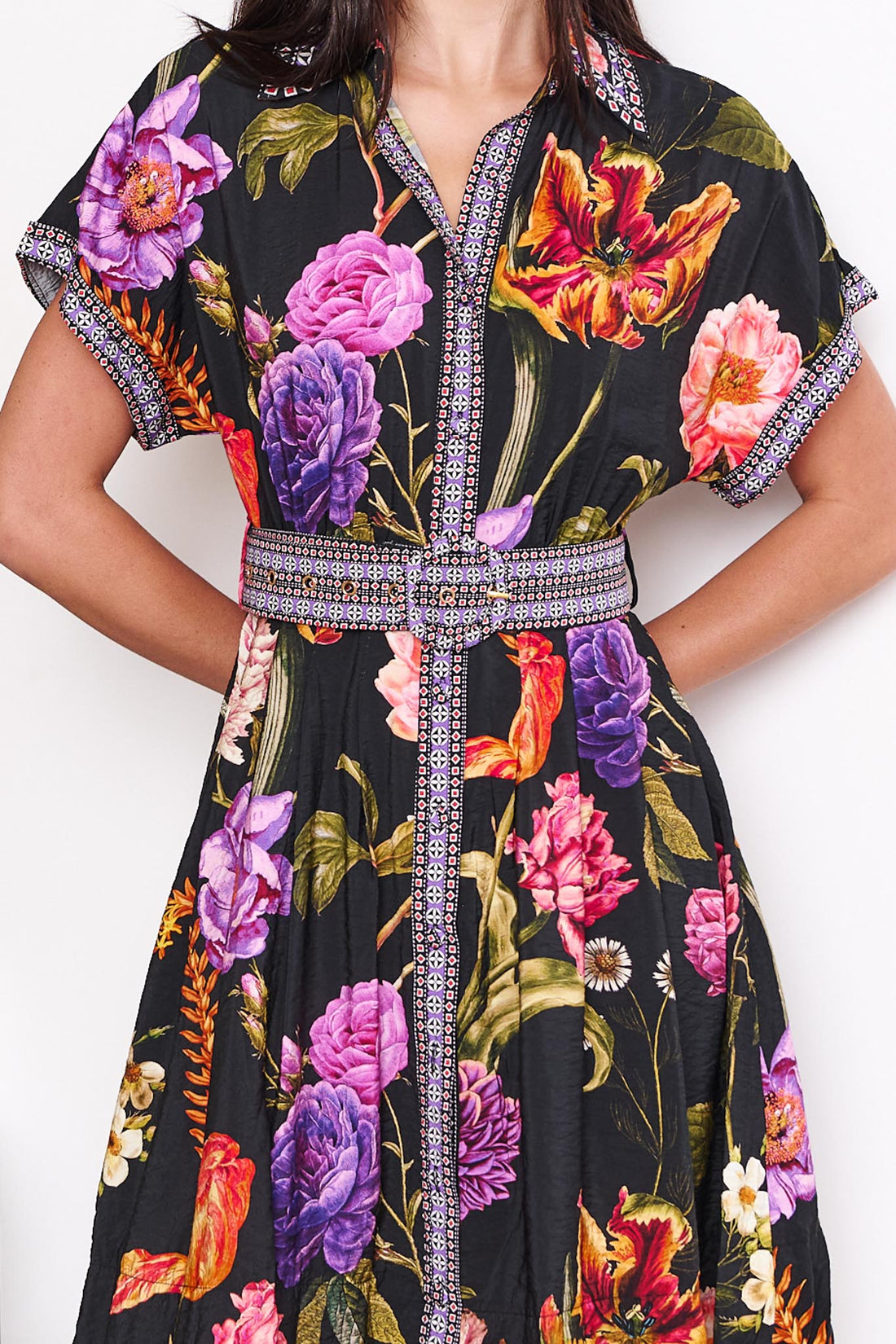 Detail of a woman wearing the minnie + MAXXIE Jemima dress with a vibrant multicolour floral print on a black background and a patterned waist tie, set against a white wall.