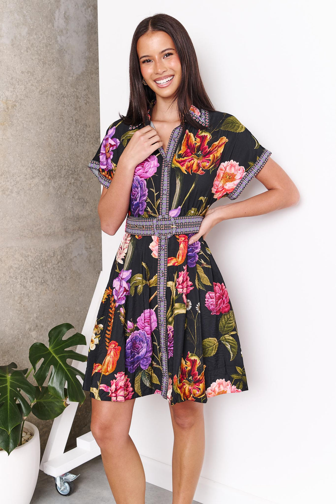 A woman wearing the minnie + MAXXIE Jemima dress with a vibrant multicolour floral print on a black background and a patterned waist tie, set against a white wall.