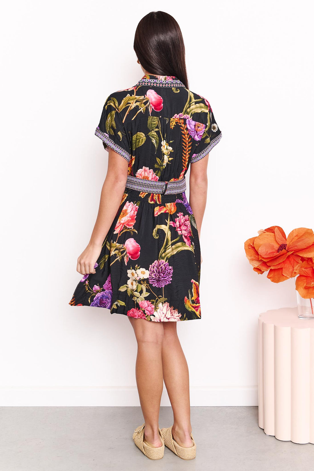 Back view of a woman wearing the minnie + MAXXIE Jemima dress with a vibrant multicolour floral print on a black background and a patterned waist tie, set against a white wall.