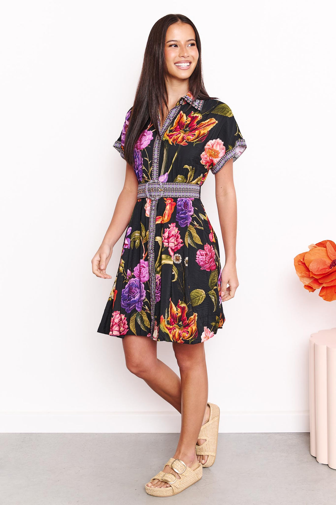 A woman wearing the minnie + MAXXIE Jemima dress with a vibrant multicolour floral print on a black background and a patterned waist tie, set against a white wall.
