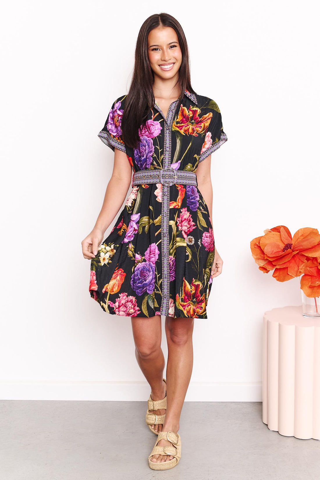 A woman wearing the minnie + MAXXIE Jemima dress with a vibrant multicolour floral print on a black background and a patterned waist tie, set against a white wall.