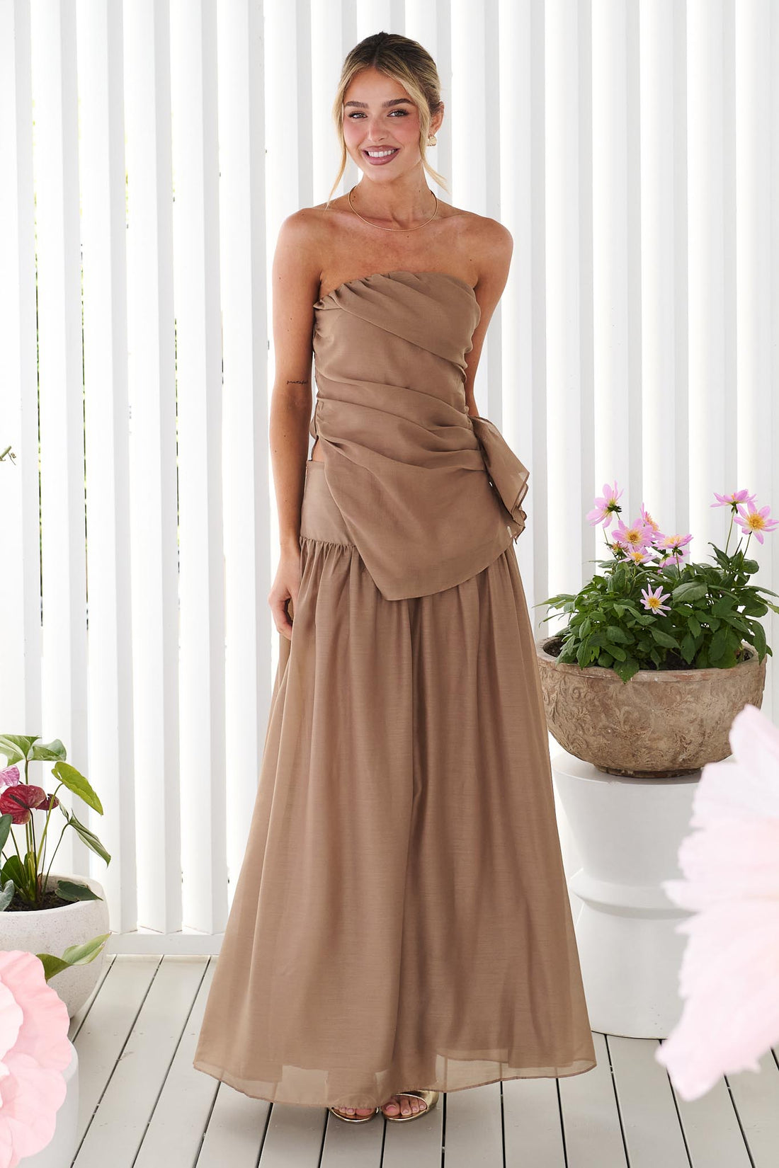 A woman wearing the minnie + MAXXIE strapless mocha brown event set with a ruched bodice and flowing maxi skirt, standing in front of a white panelled wall.
