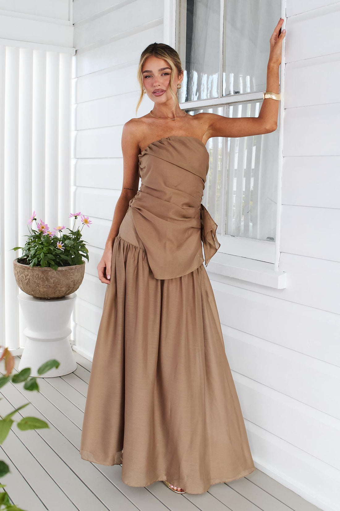 A woman wearing the minnie + MAXXIE strapless mocha brown event set with a ruched bodice and flowing maxi skirt, standing in front of a white window.
